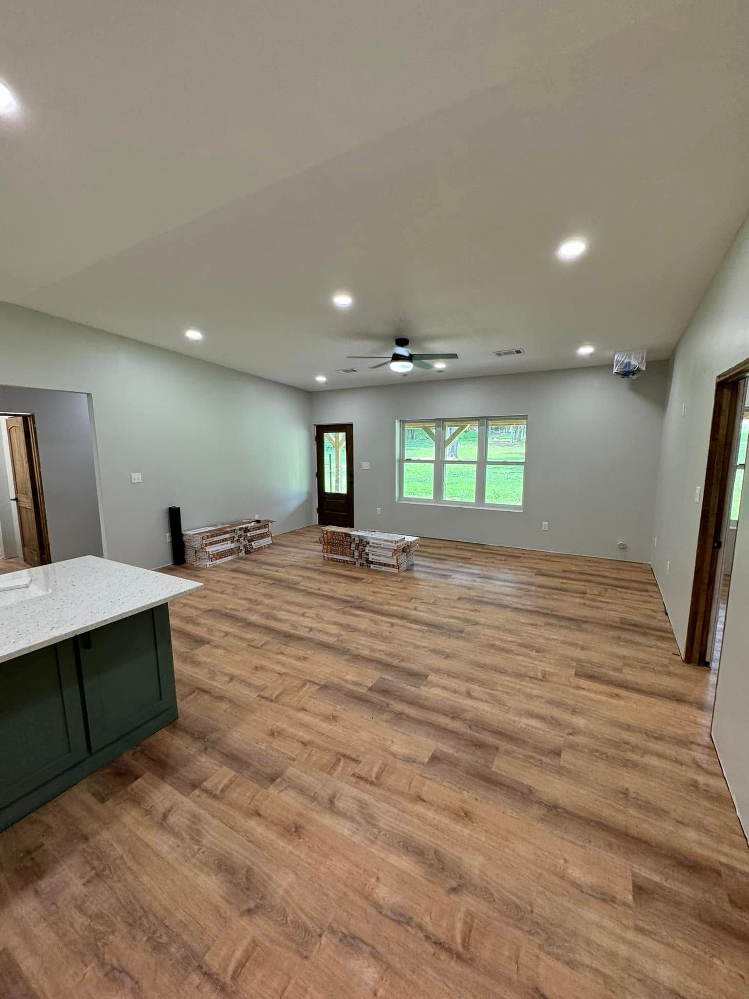 Spacious, newly constructed living room with wood-look flooring, green cabinets, and recessed lighting. A dark exterior door and window with a view are visible.