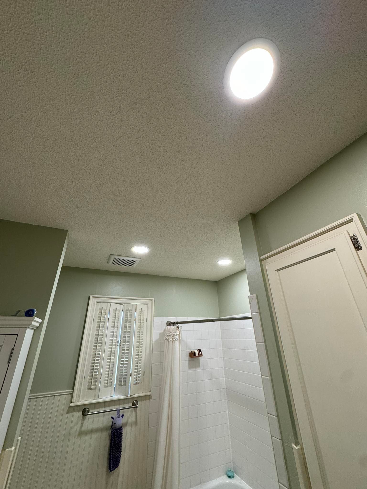Bathroom ceiling with recessed lights and green walls. White shower curtain, window with a patterned curtain, and a closed white door are also visible.