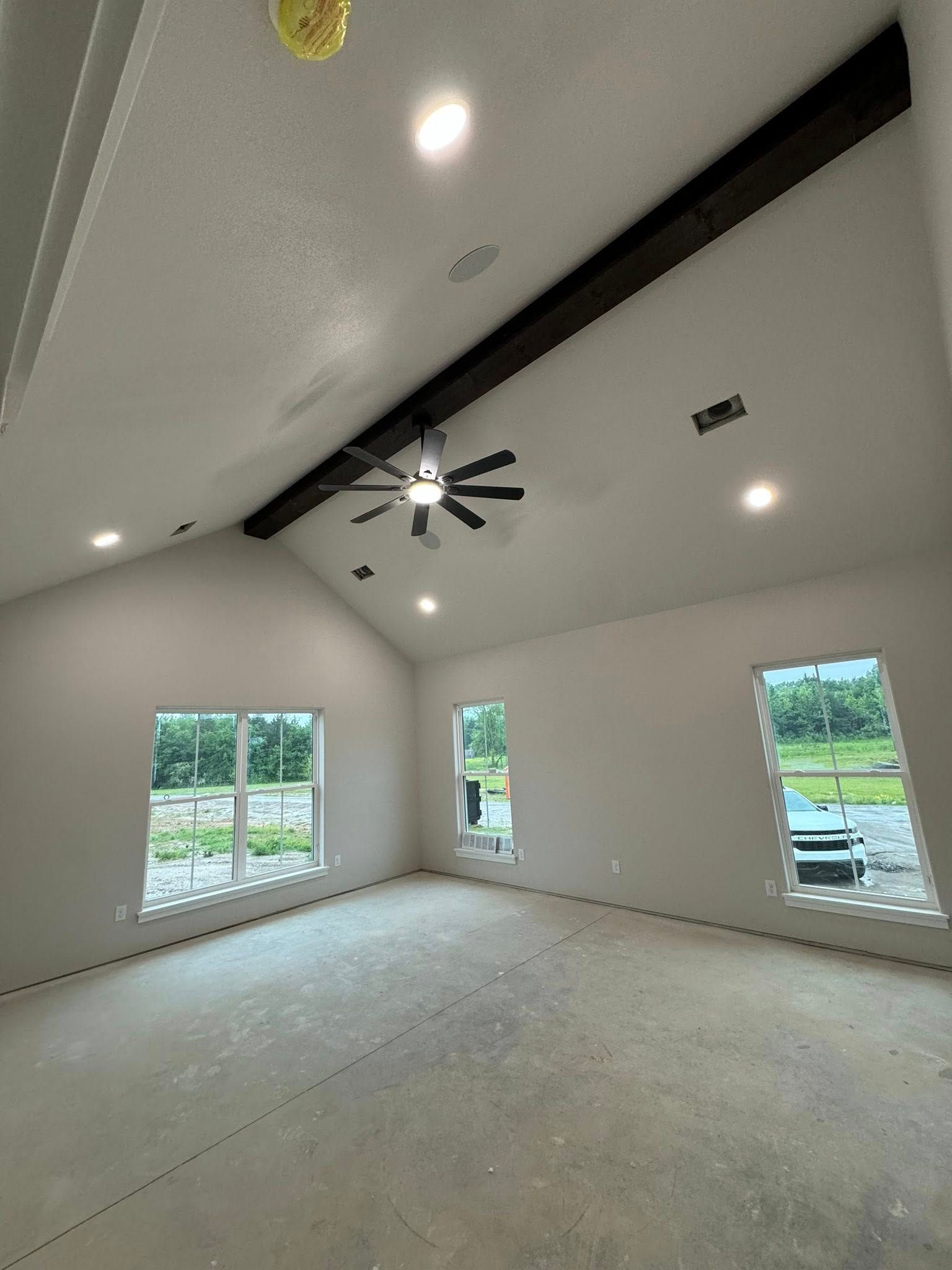 An empty room with a high, angled ceiling and recessed lighting, with a dark wooden beam and ceiling fan. Three windows offer views of the outdoors.