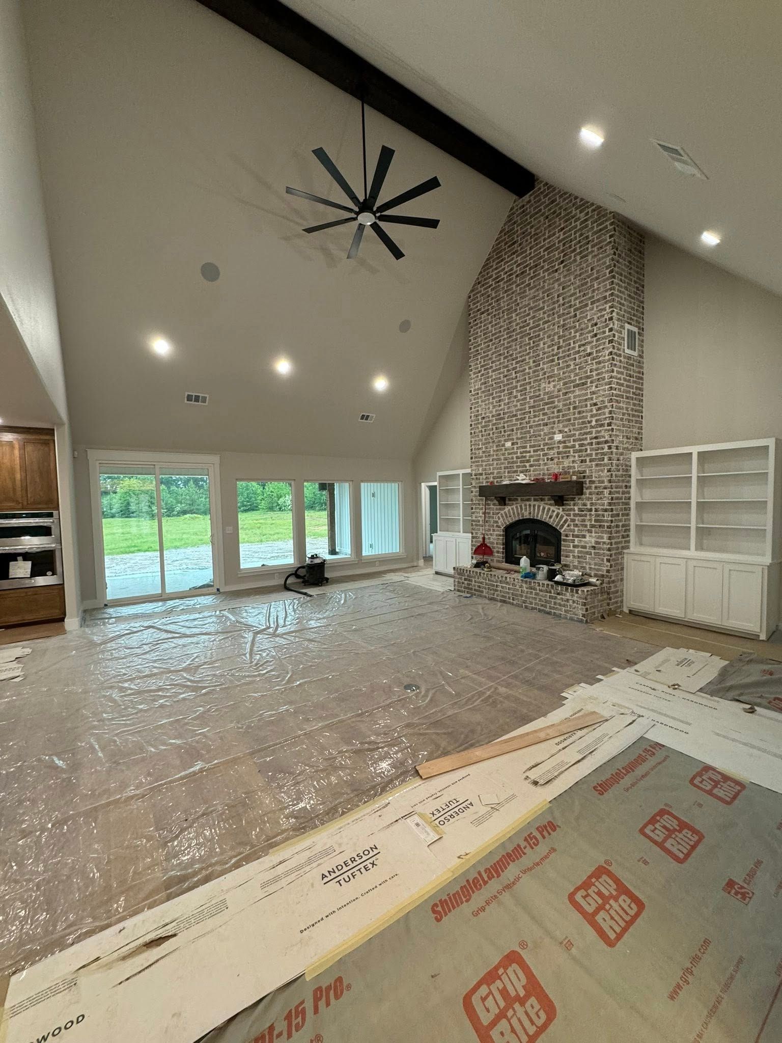 A large living room under renovation. It features a brick fireplace, tall windows, and protective floor covering.