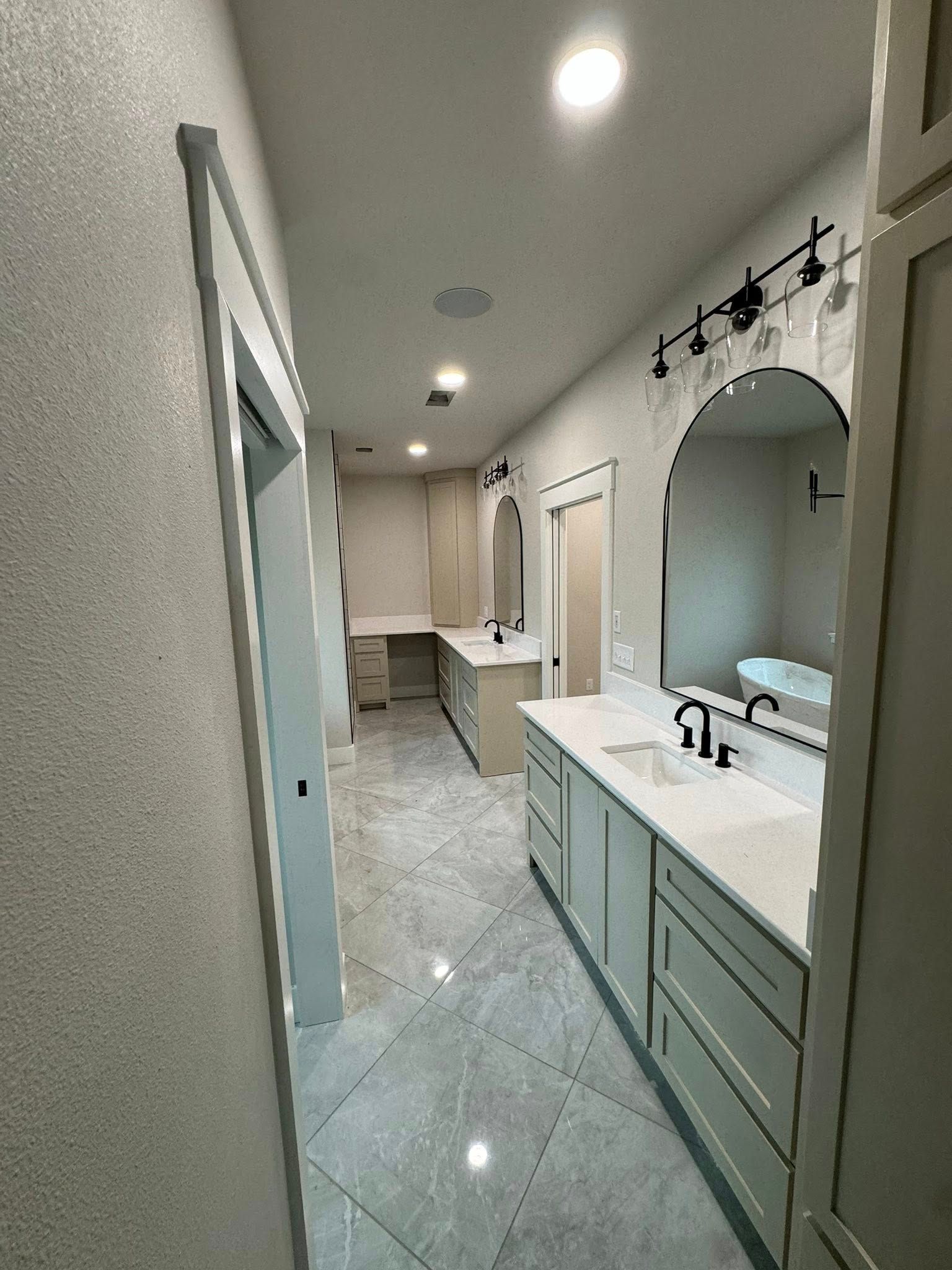 A long, modern bathroom with light gray cabinets, countertops, and tile flooring. A vanity and large mirror are visible.