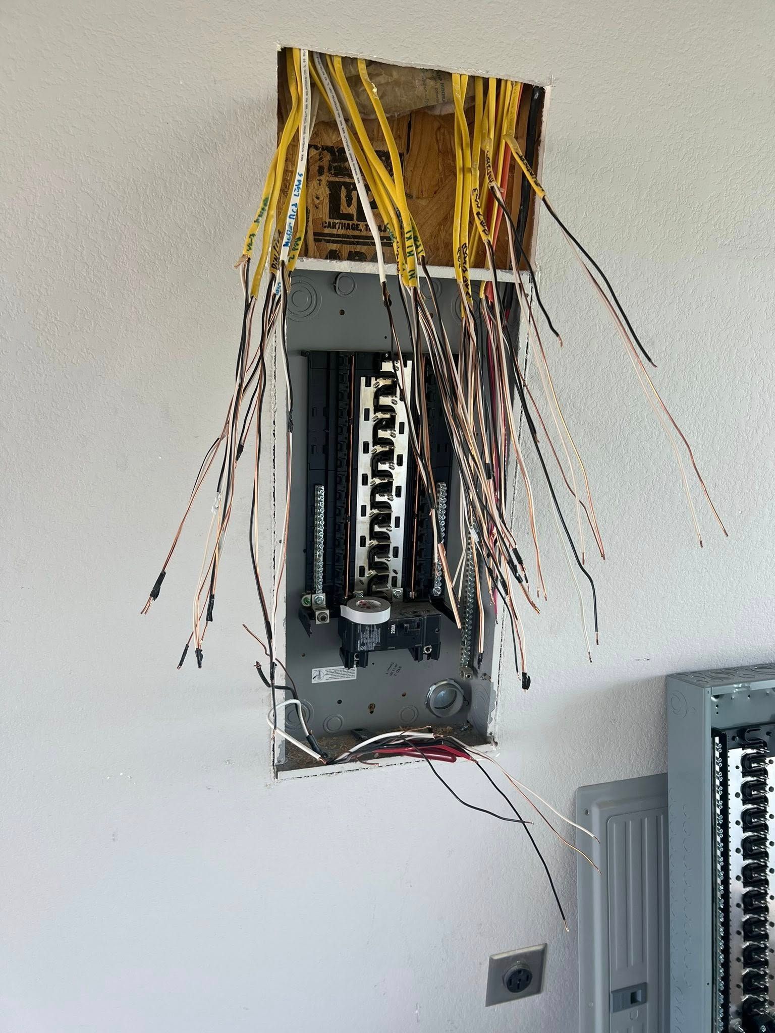 Electrical panel with exposed wires, recessed in a white wall. Wires of various colors dangle.