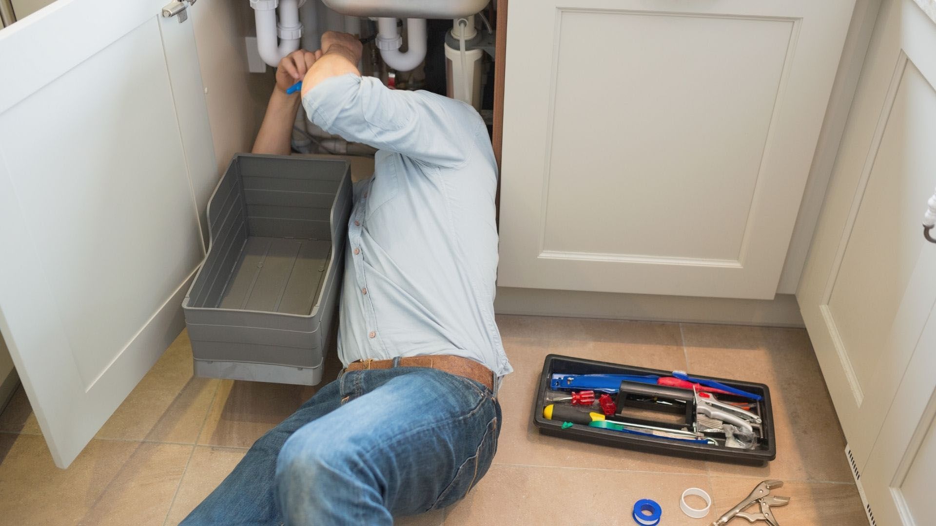 A man is kneeling on the floor under a sink in a kitchen.