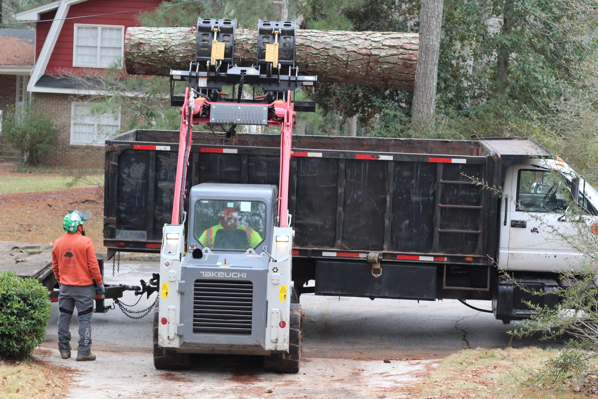 A skid steer loading a large log into a dump truck. A worker watches. Outdoors, residential setting.