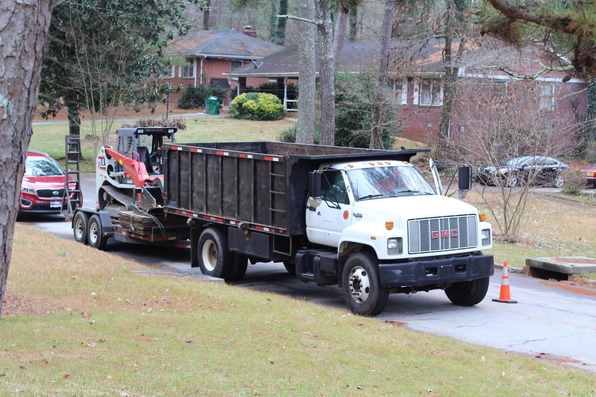 White dump truck with trailer carrying a skid steer on a residential street.