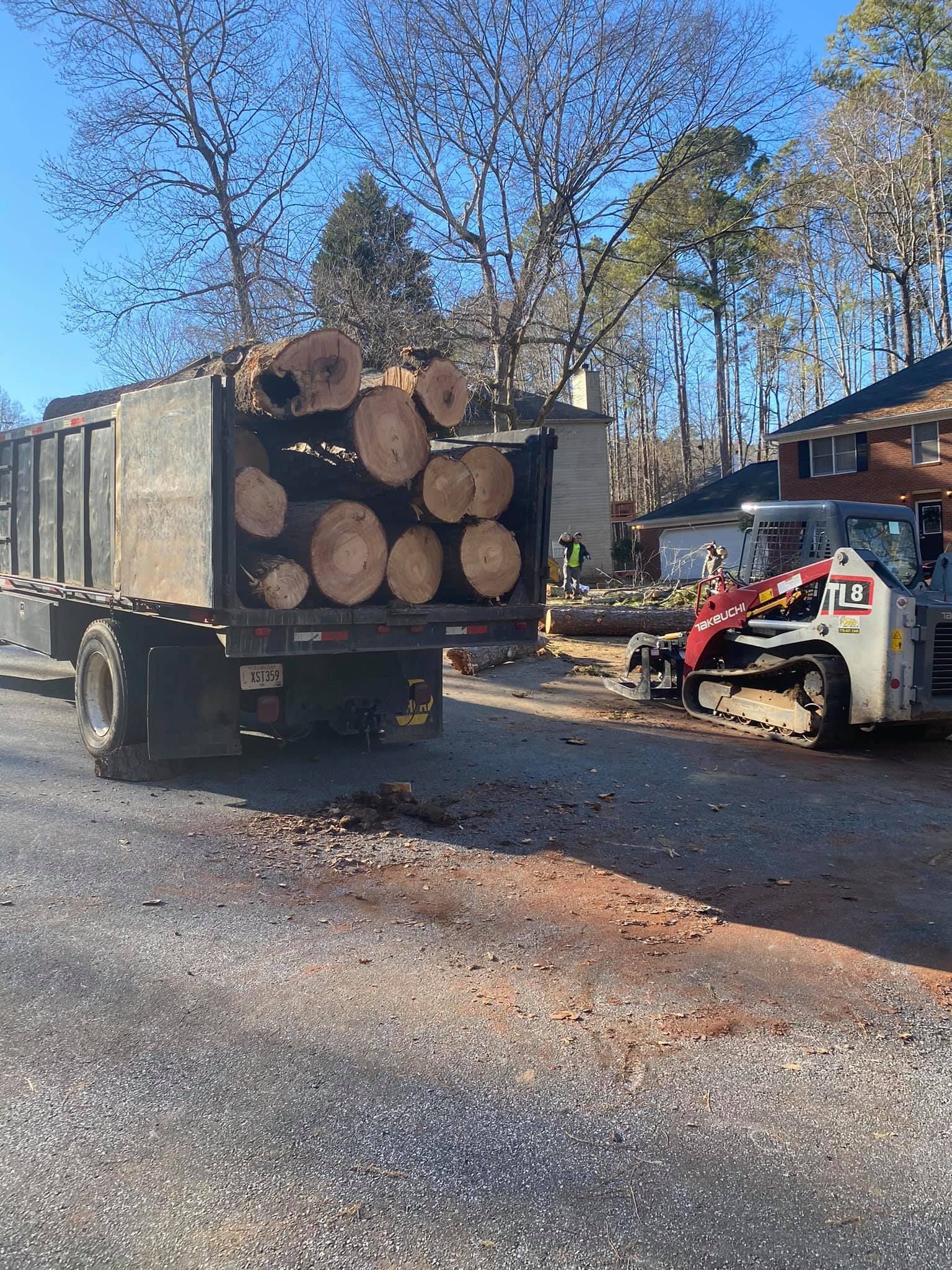 Truck loaded with logs, a small tractor nearby; trees and a house in the background.