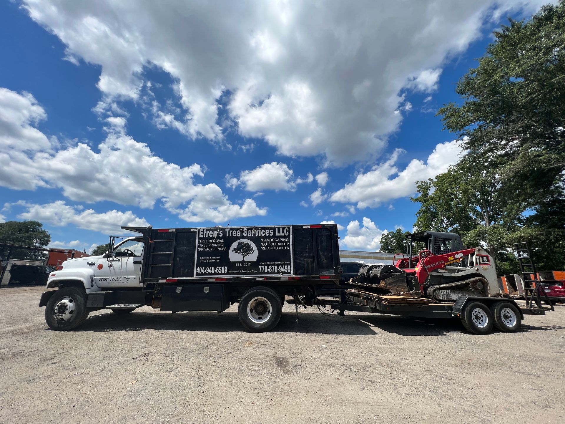 White work truck with trailer carrying a mini-excavator under a bright blue sky with puffy clouds.