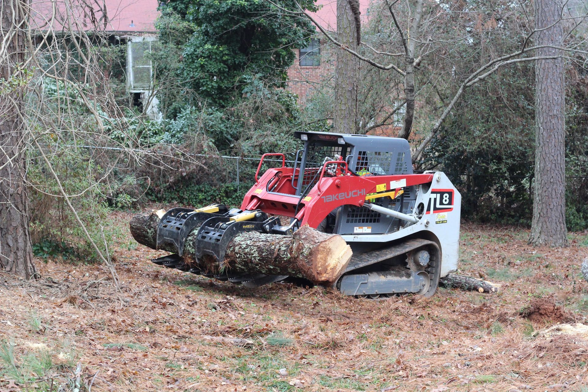 Skid steer loader with log grapple holding a tree trunk, in a wooded area with a house in the background.