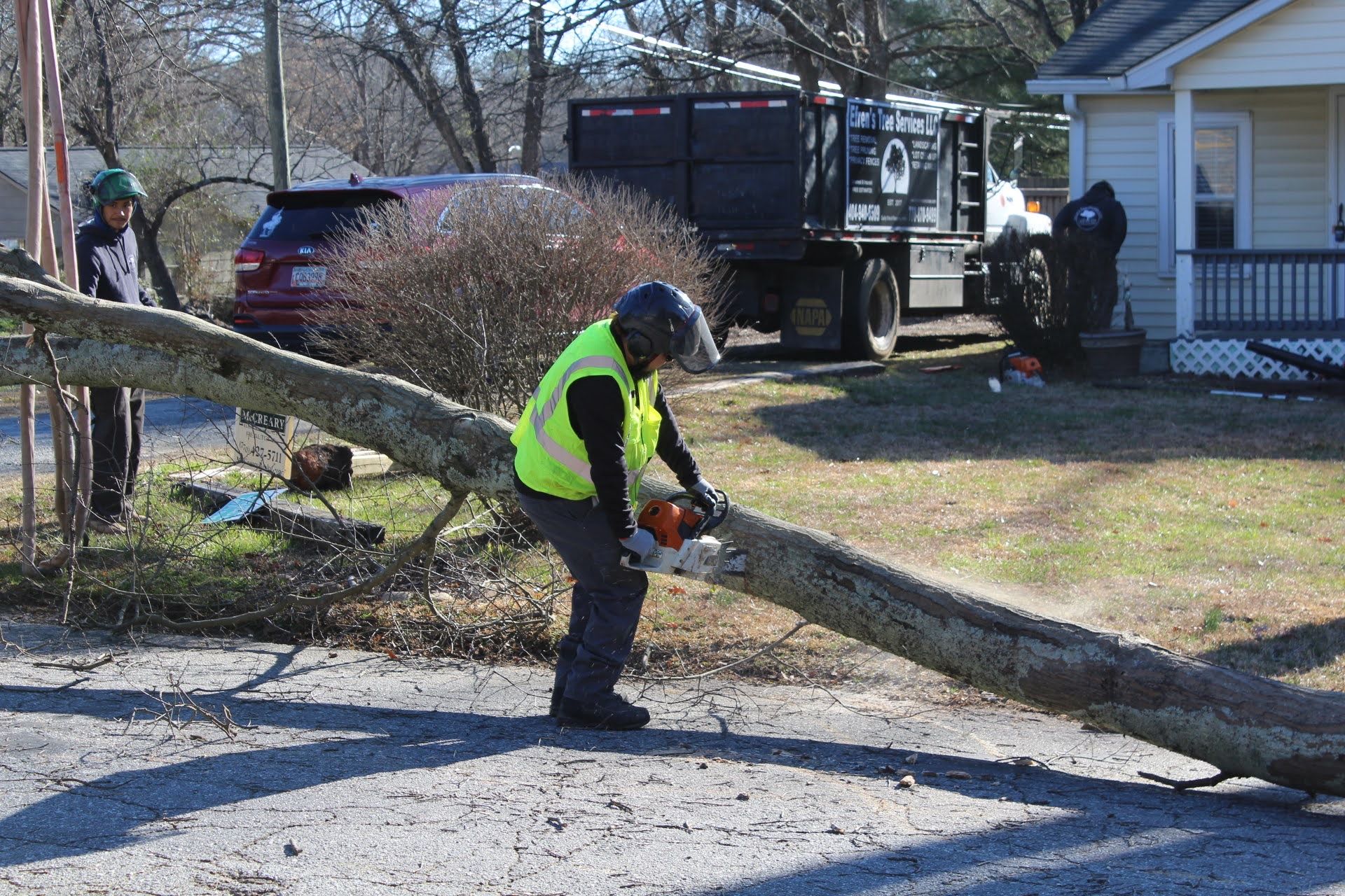 Worker in safety vest uses chainsaw on fallen tree in front of a house, other workers nearby.