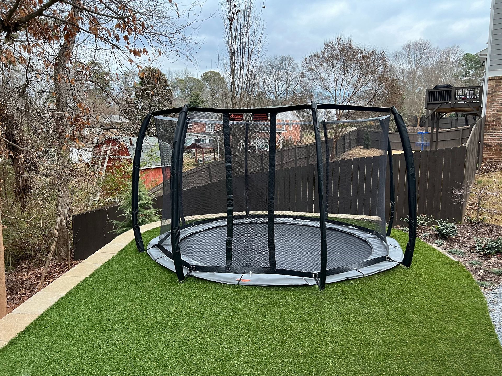 Black trampoline with safety net on green artificial turf. Outdoor setting with trees and fence.