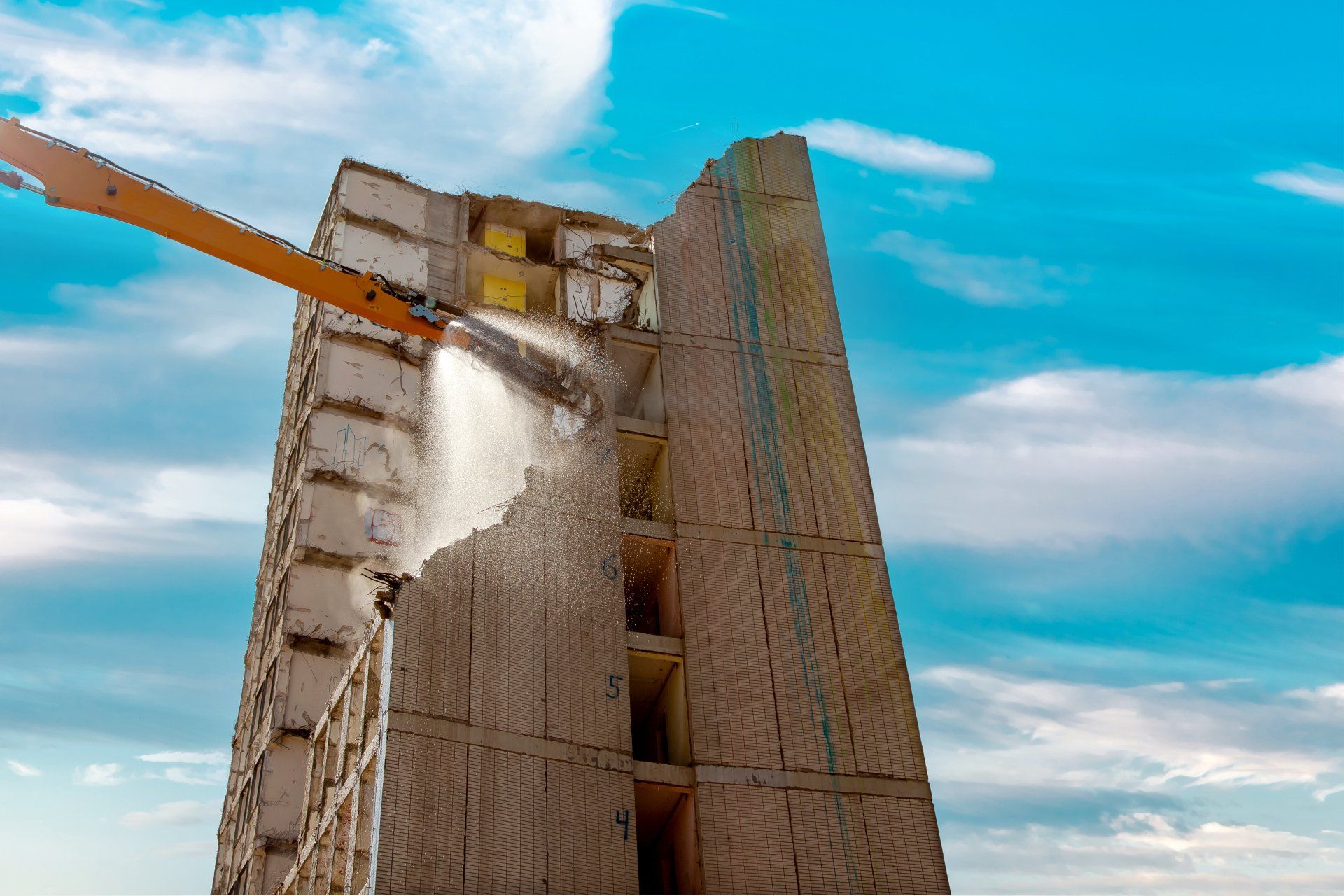 Image of a building being demolished by a crane that simultaneously sprays it with water to reduce dust and debris from becoming airborne.
