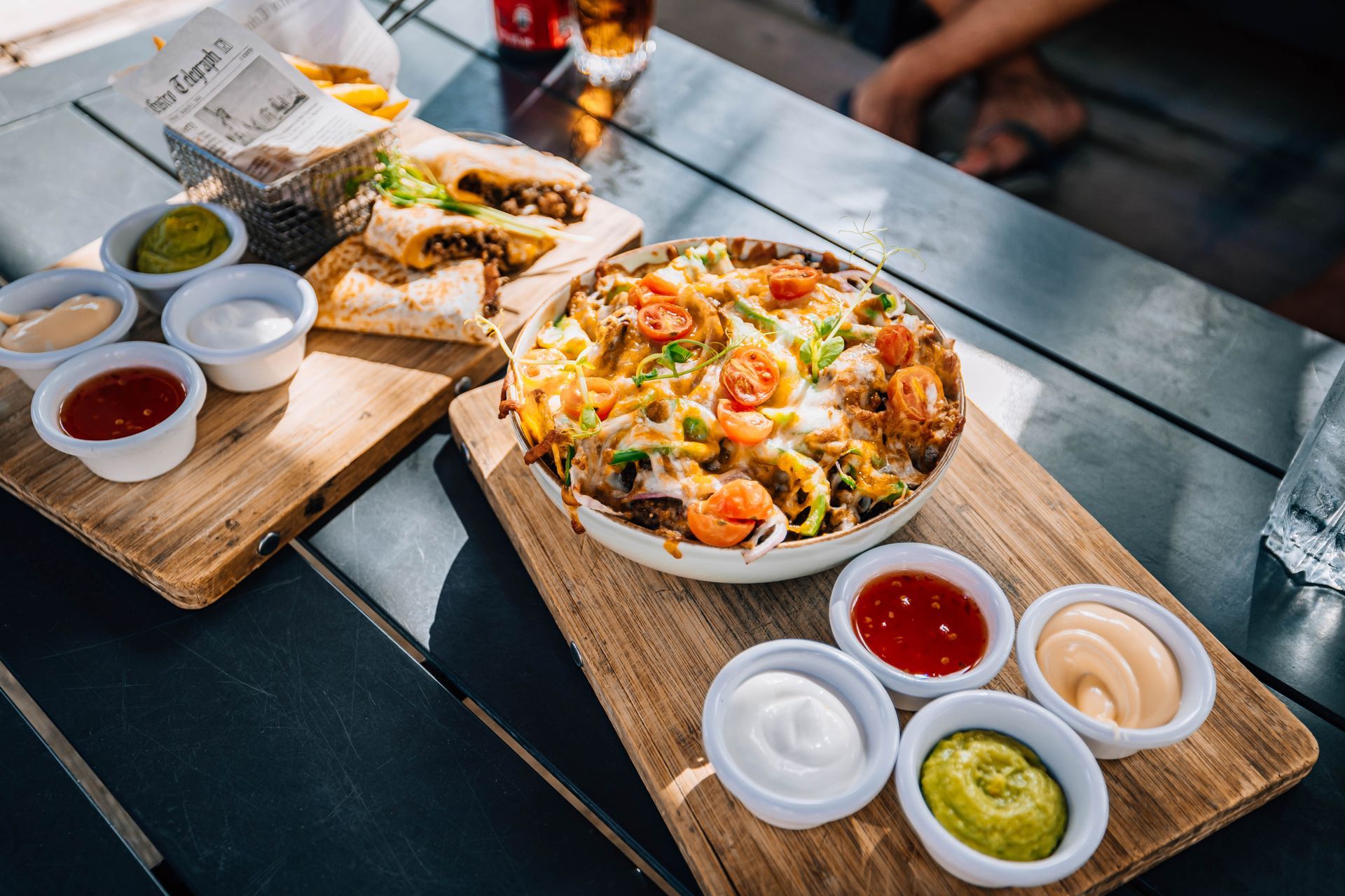 Two wooden boards on a table holding a loaded skillet of cheesy nachos, quesadillas, and bowls of dipping sauces.