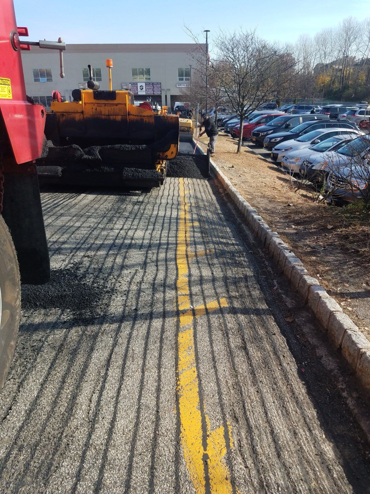 Asphalt paving a parking lane. A paving machine lays fresh asphalt over a yellow line next to a curb and parked cars.