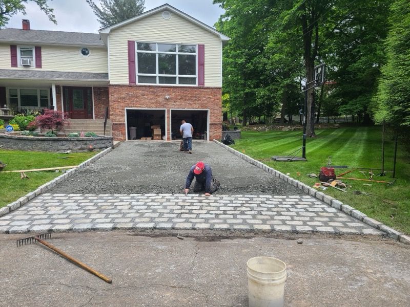 Construction workers laying paving stones on a driveway. House with a two-car garage in background. Green grass on either side.