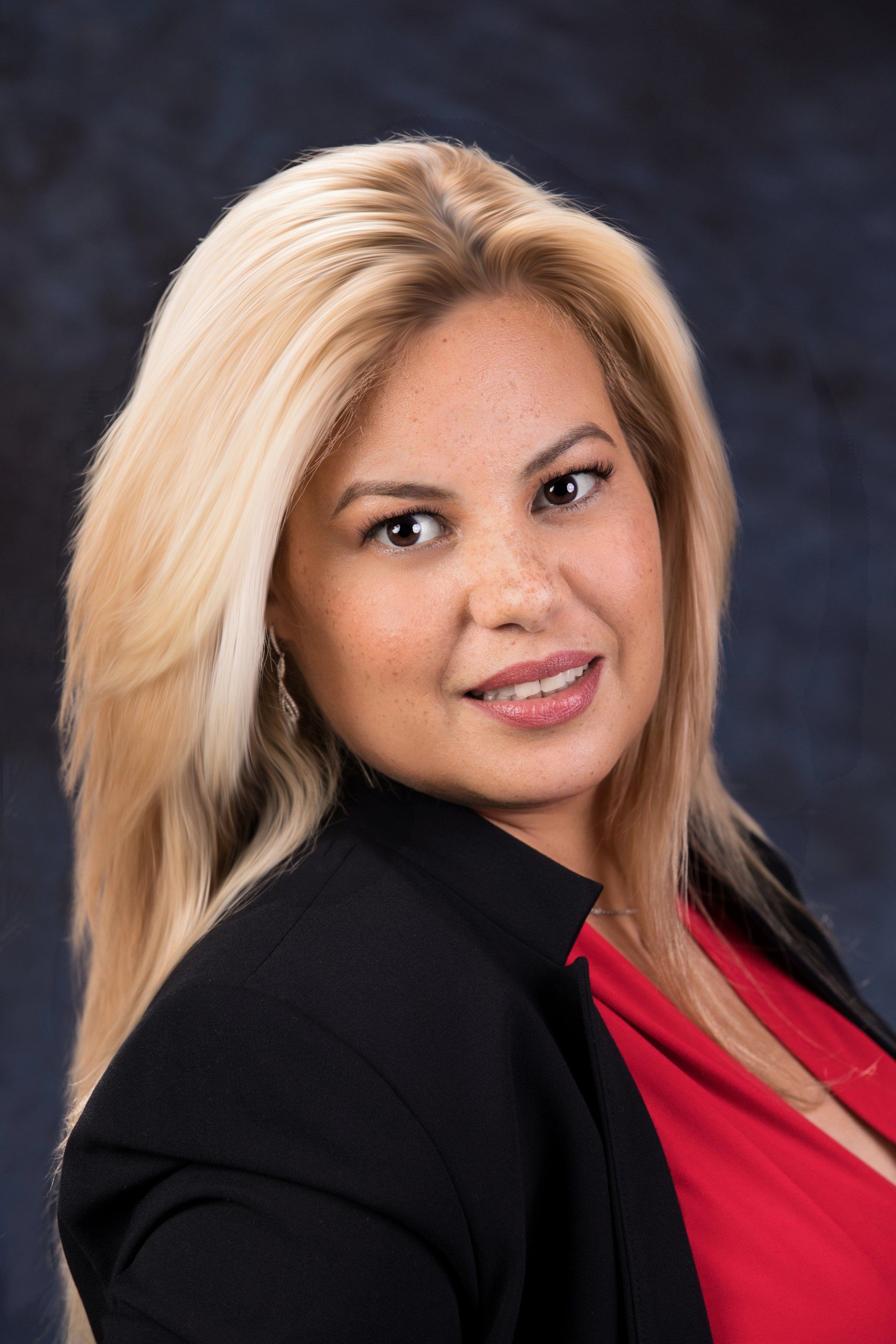 A woman with blonde hair, wearing a black blazer over a red top, poses against a dark, textured background.