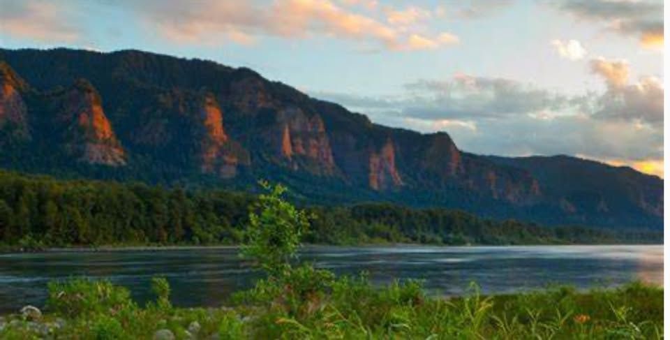 River flows along dark mountain range, soft sunlight highlights cliffs, green foliage in foreground.