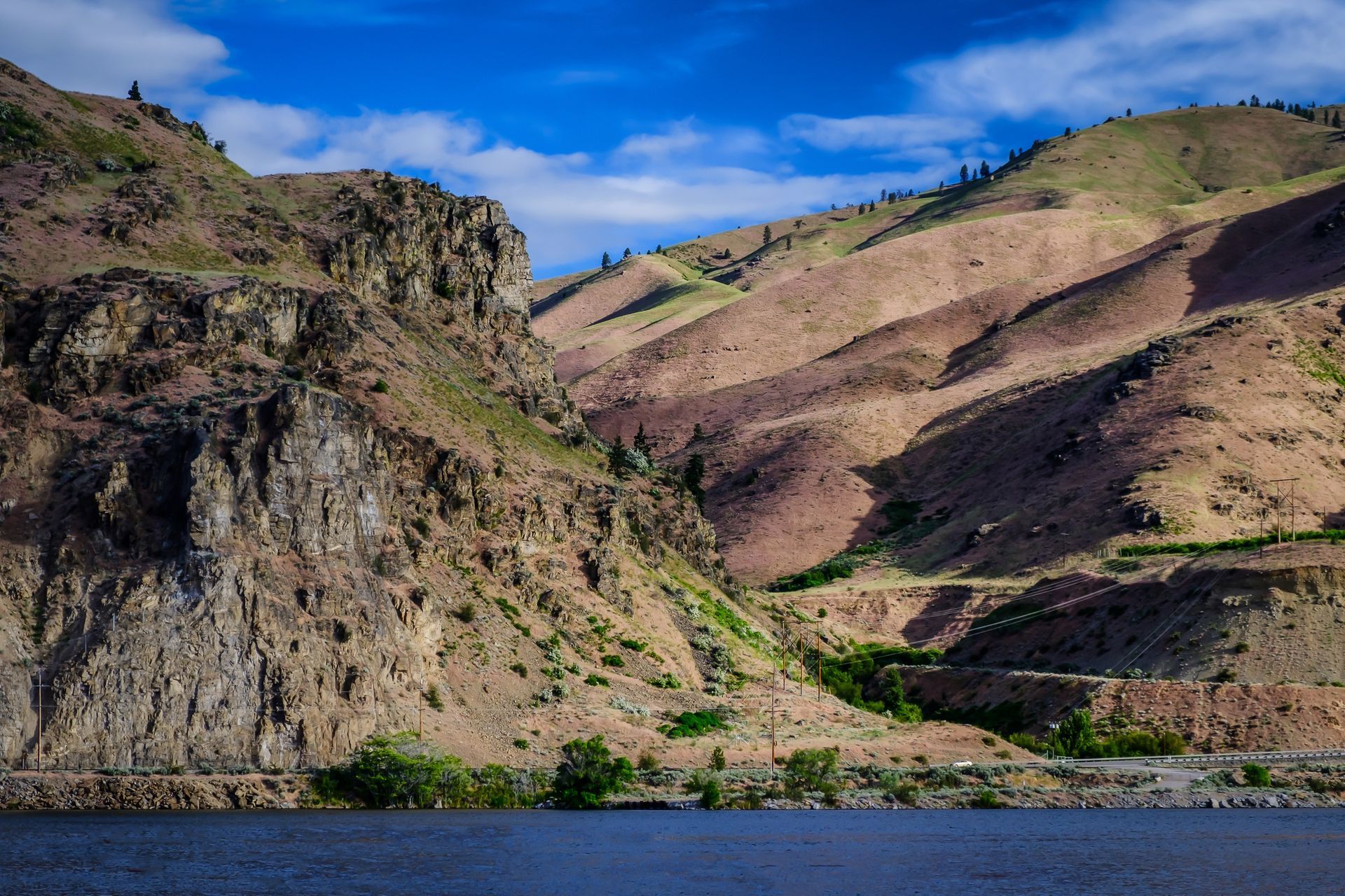 Rocky cliffs and rolling, brown hills rise above a dark blue lake under a partly cloudy sky.