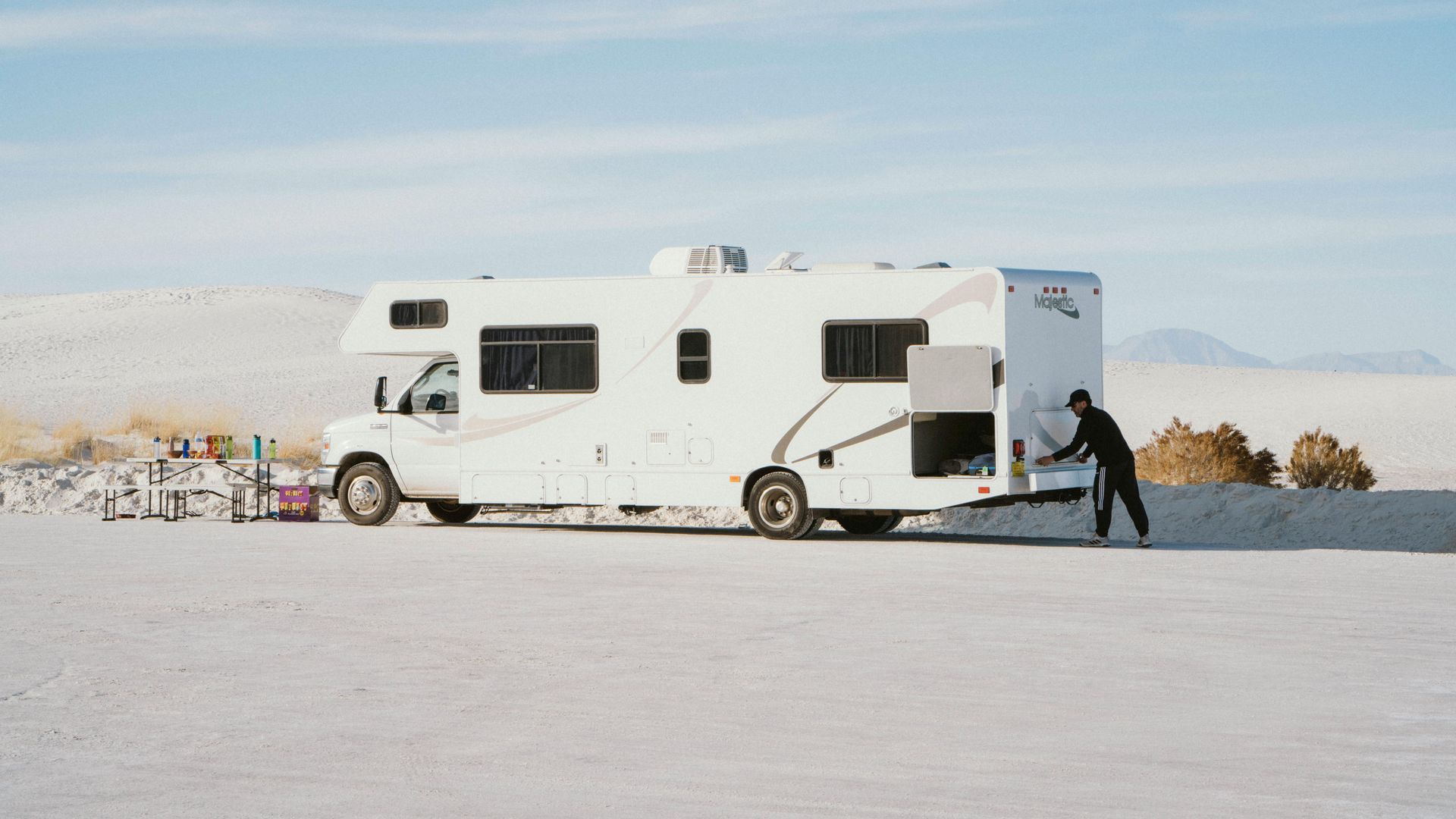 White RV parked in a bright desert landscape; person near back opening compartment, skis leaning nearby.