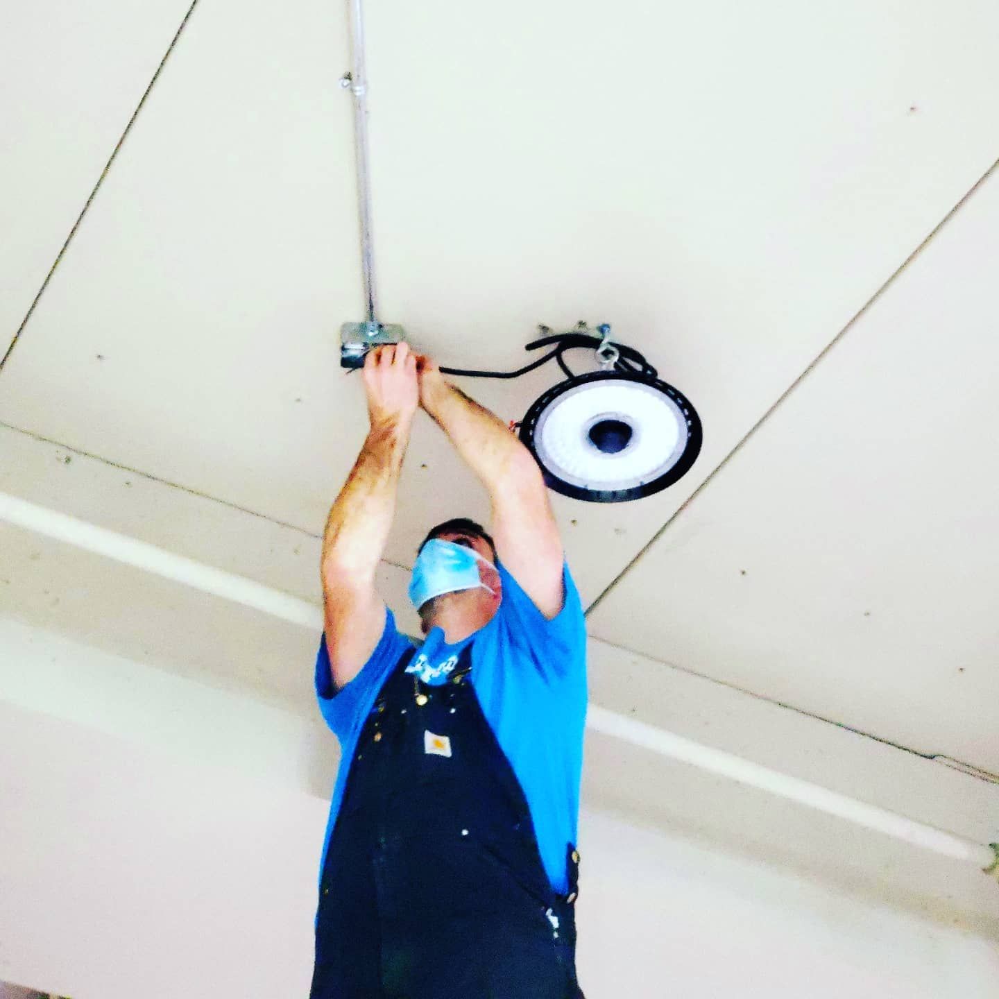 A man wearing a blue shirt and black overalls is working on a ceiling light