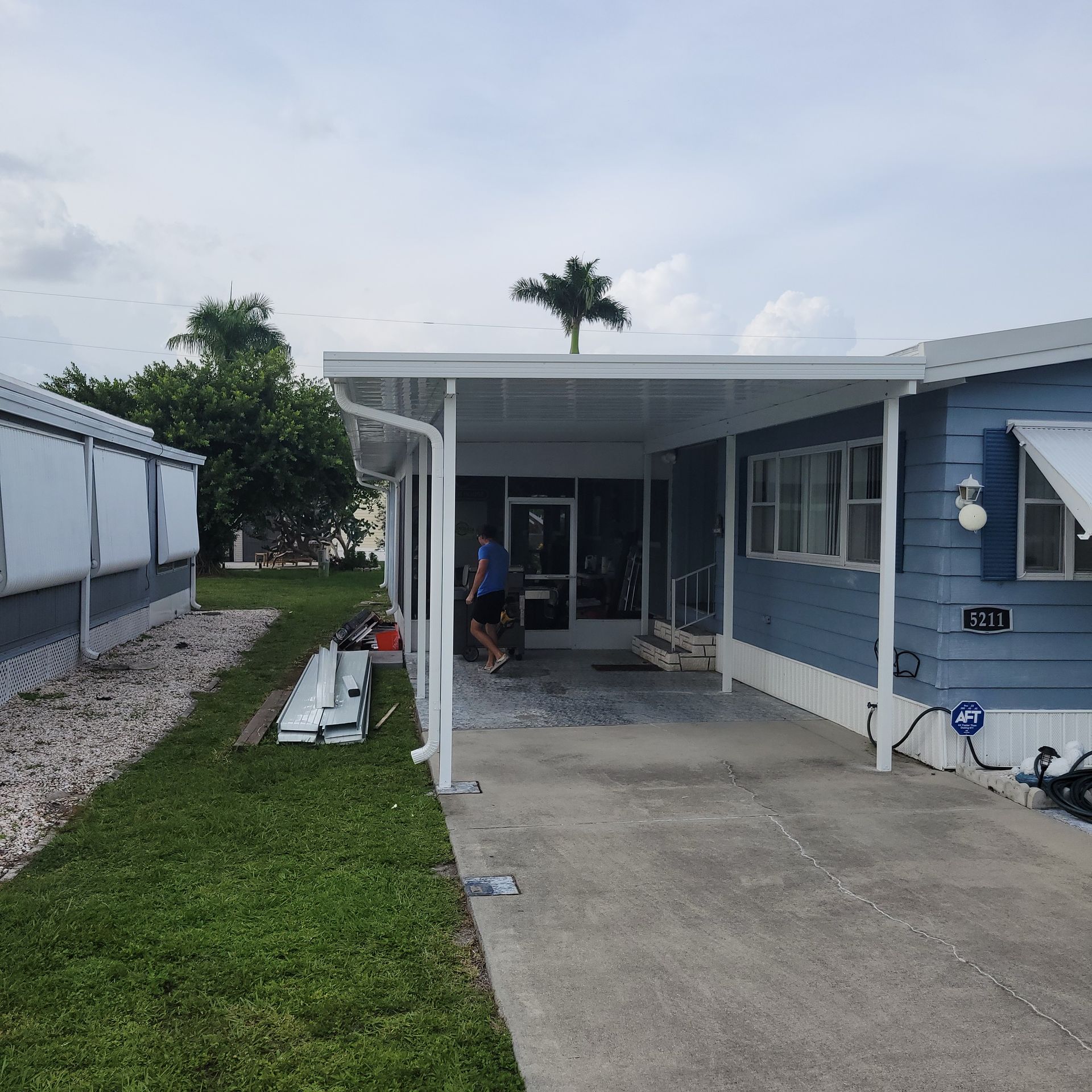 A blue mobile home with a covered driveway and a palm tree in the background