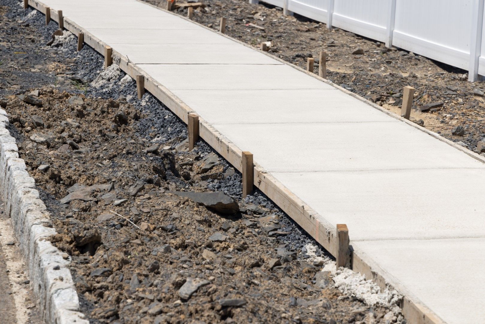 Concrete sidewalk under construction, bordered by wooden forms and dirt, next to a white fence.