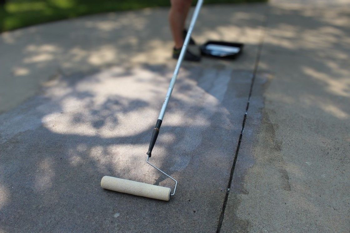 Person using a paint roller to seal a concrete driveway with a paint tray in the background.
