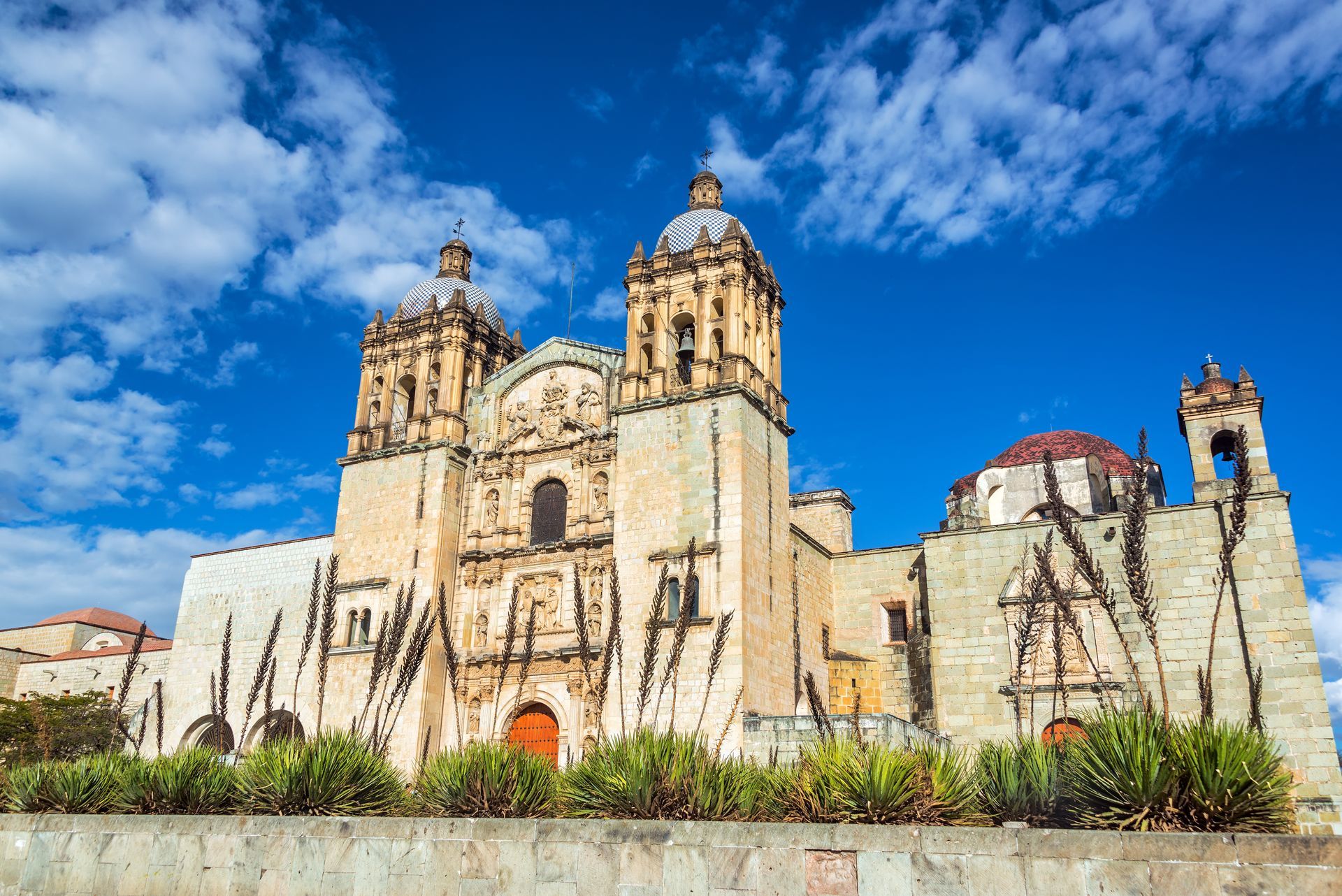 A historic stone church with two bell towers, a red-tiled dome, and small green plants in front under a blue sky.