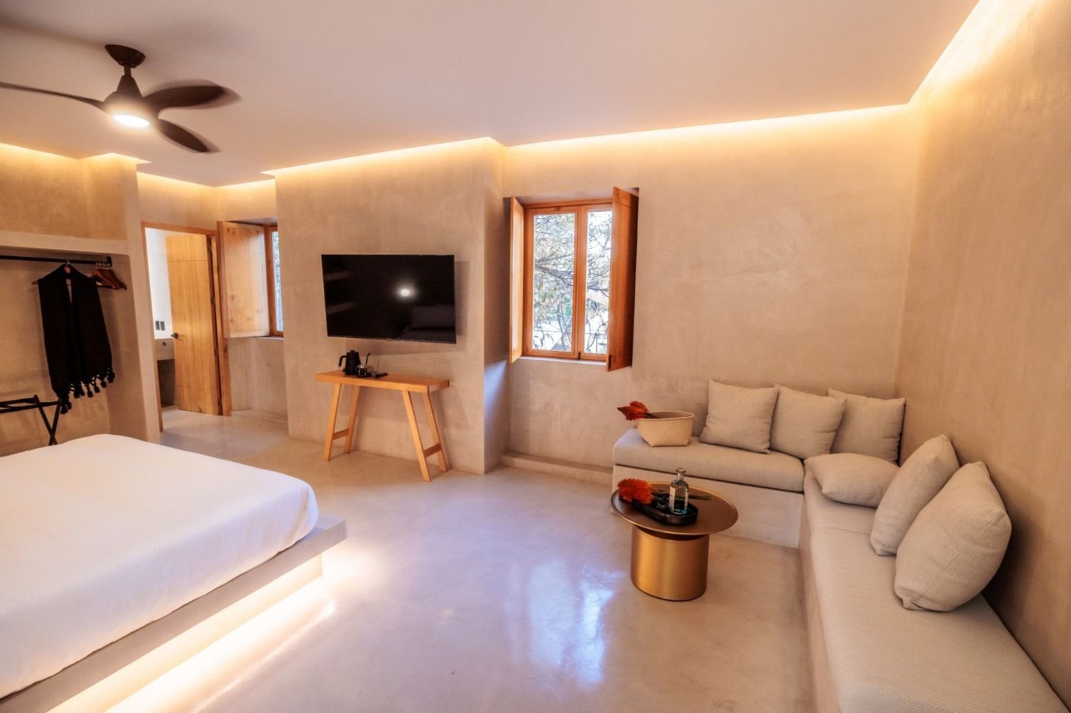 A modern, neutral-toned hotel room with a bed, a TV on a wooden stand, a window, and a built-in sofa with a coffee table.