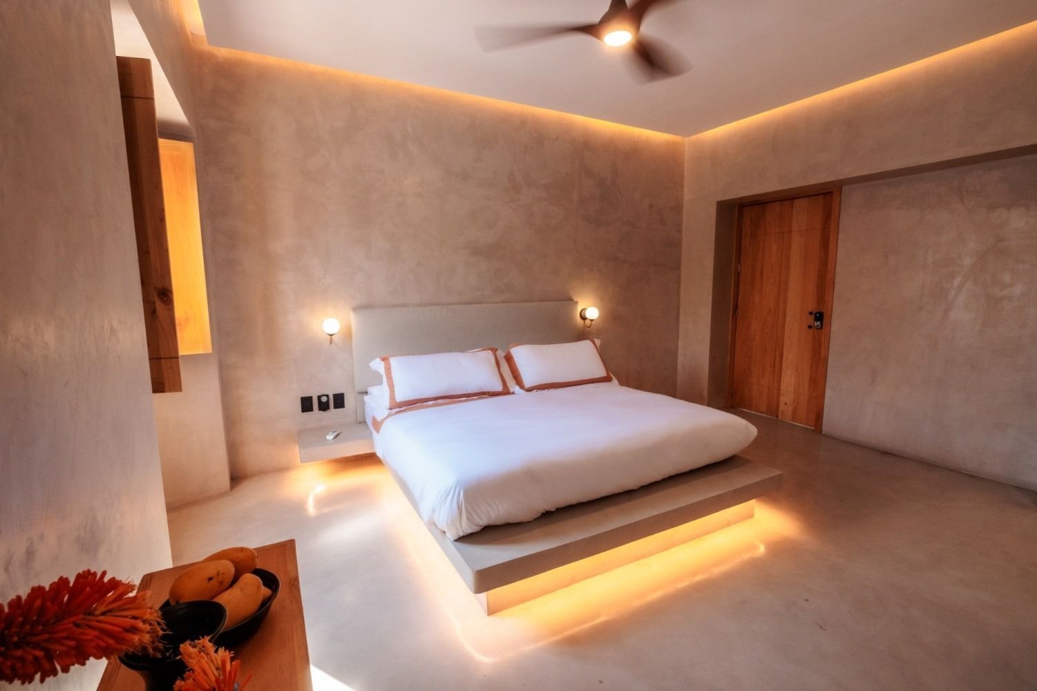 A modern, minimalist bedroom with warm cove lighting under a platform bed, white bedding, and smooth, sandy-toned walls.