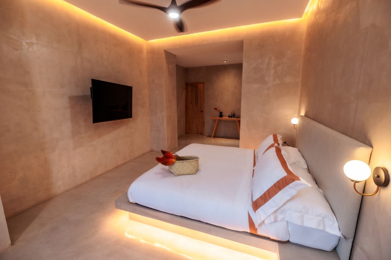 A modern, minimalist bedroom with beige plastered walls, a platform bed with white linens, warm lighting, and a TV.