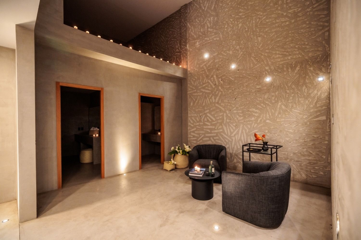 A modern lounge with grey walls, two dark armchairs, a small round table, and two doorways, lit by recessed ceiling lights.