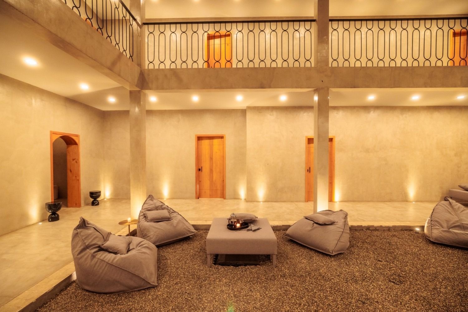 A minimalist lounge with gravel flooring, bean bag chairs, a low central table, and warm wall lighting in a large room.
