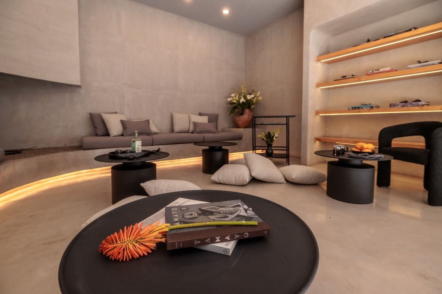 A modern, neutral-toned lounge with built-in seating, low tables, floor cushions, and shelves with decorative items.