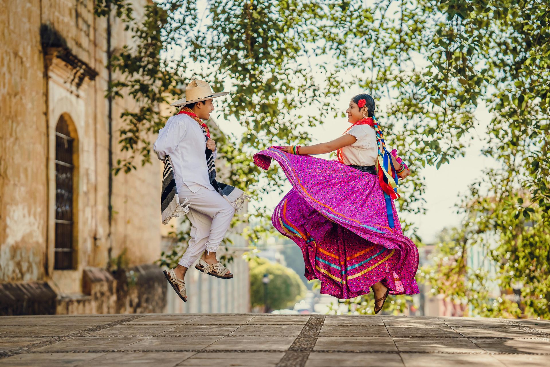 Dancers in traditional Mexican attire jump mid-air on a stone path next to a colonial-style building and lush trees.
