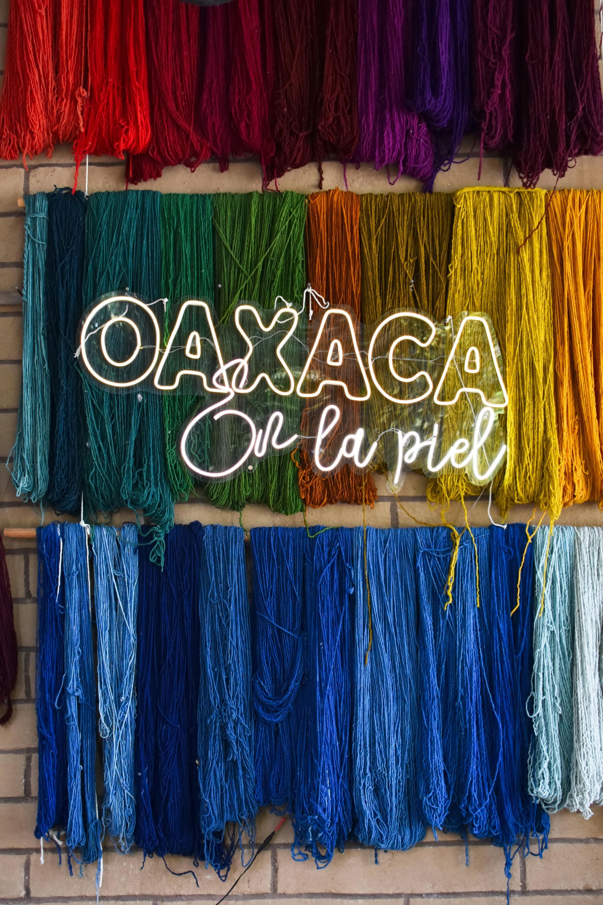Rows of vibrant, multicolored yarn hang against a brick wall, with a neon sign reading