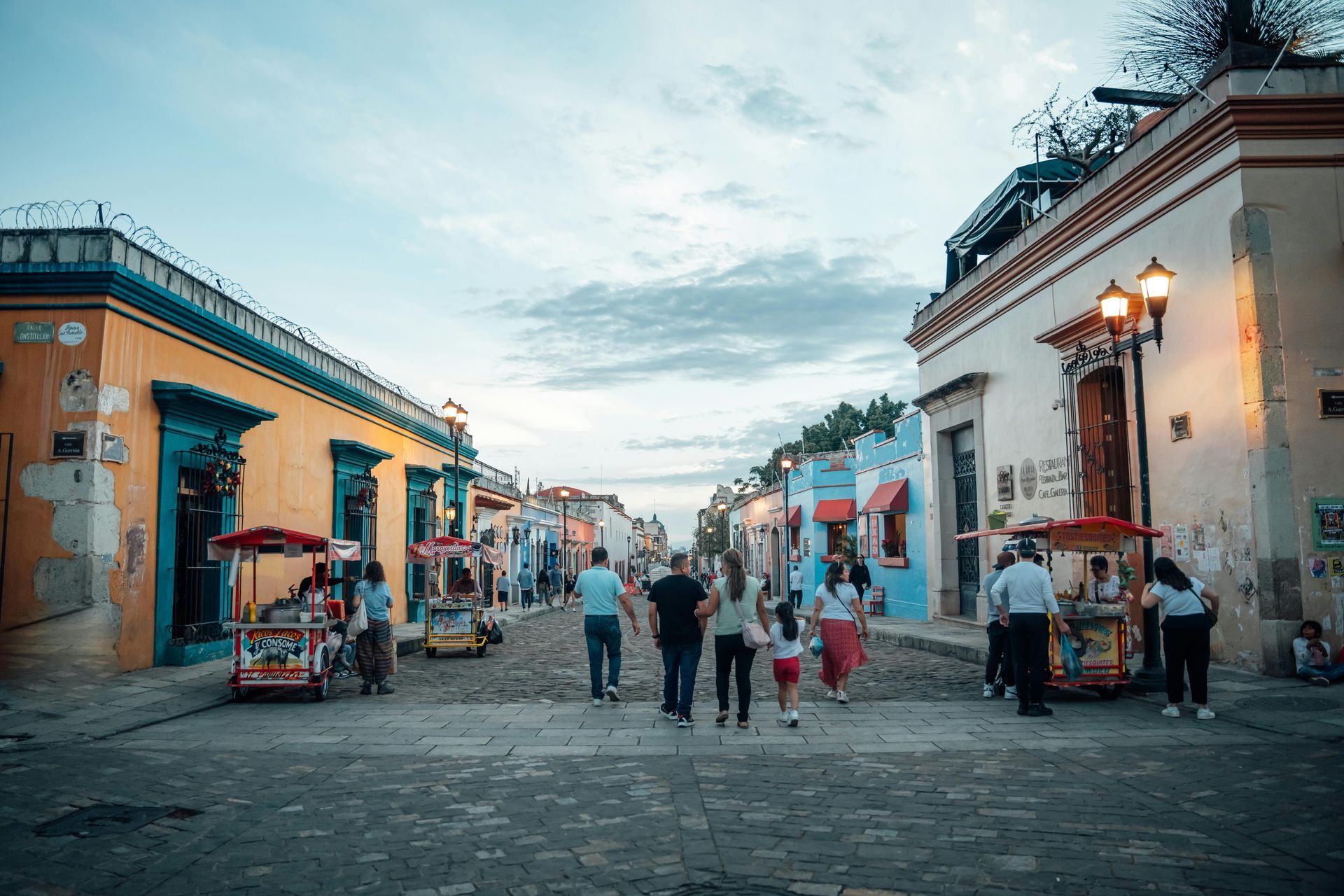 A cobblestone street in a colorful colonial town with people walking and street vendors with carts.