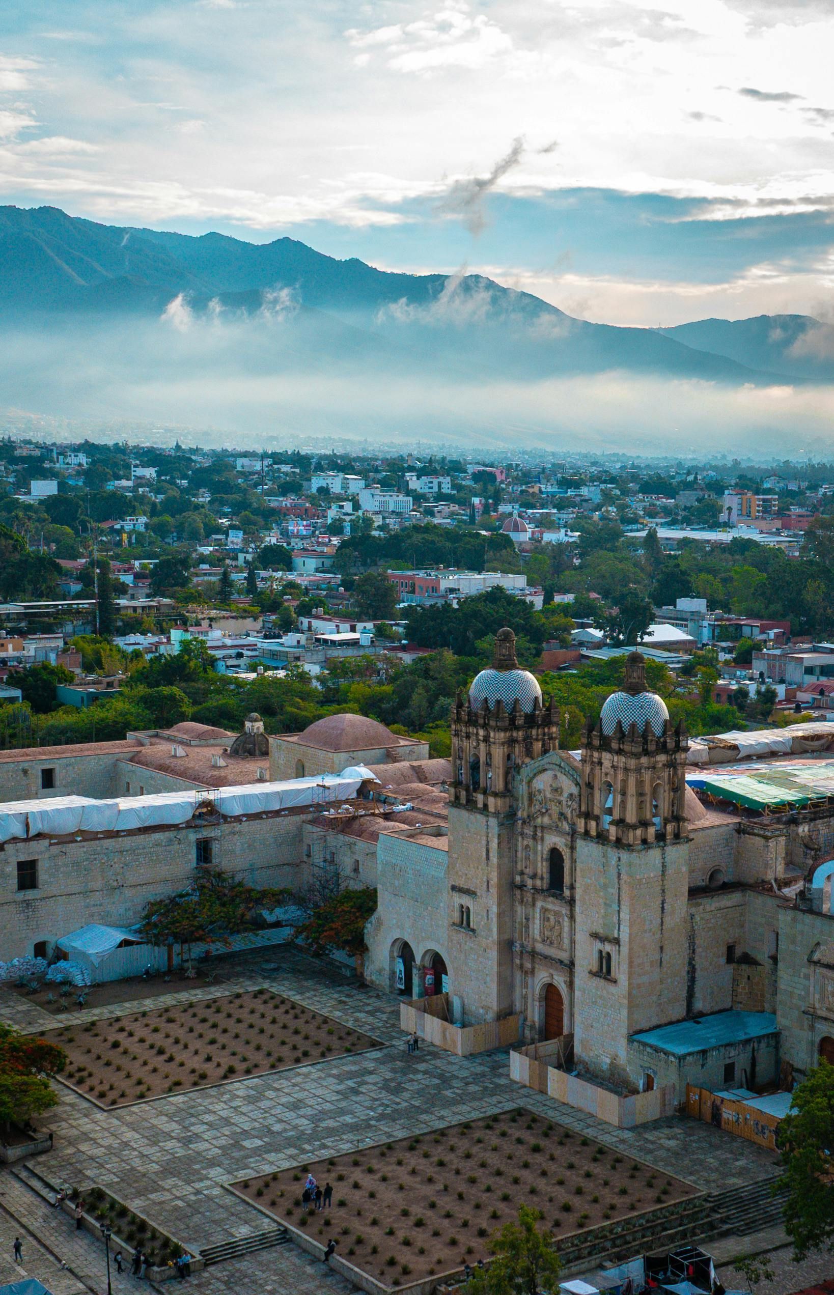 Aerial view of the Santo Domingo Church in Oaxaca, Mexico, with tiled courtyards and misty mountains in the background.