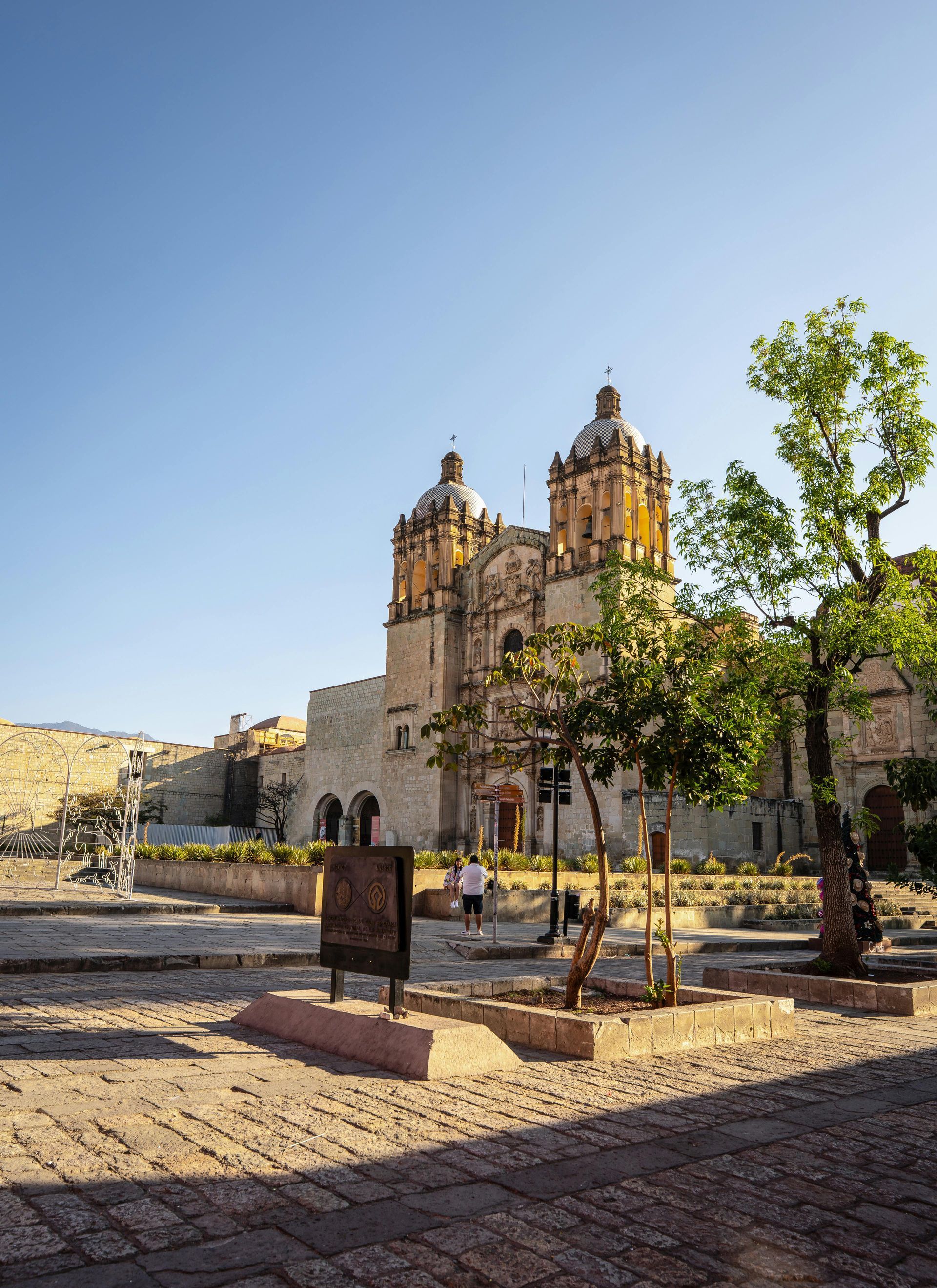A stone cathedral with two domed towers stands behind a paved plaza with trees under a clear blue sky.