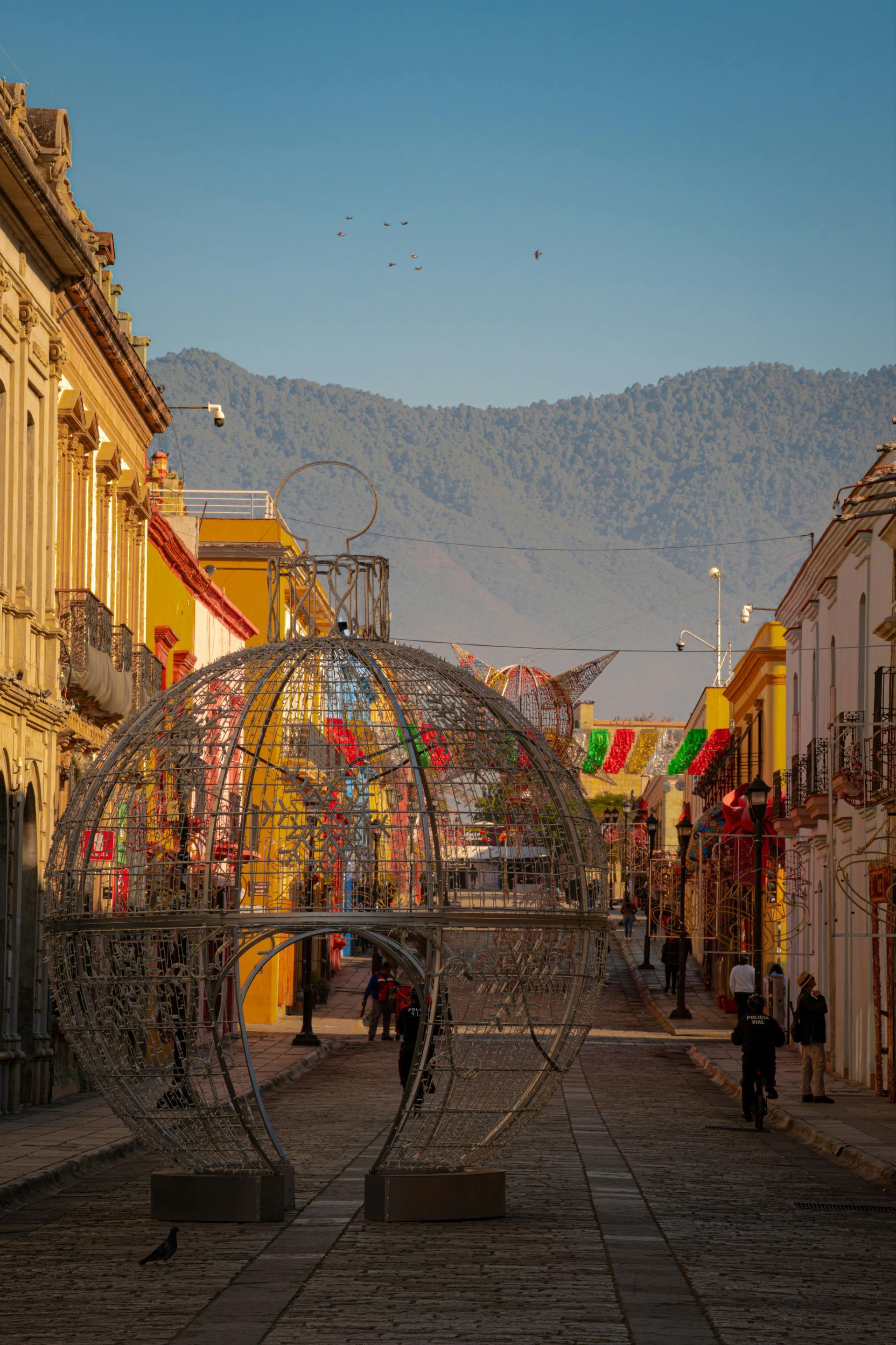 A large, spherical wire Christmas ornament stands on a cobblestone street between yellow buildings, with mountains beyond.