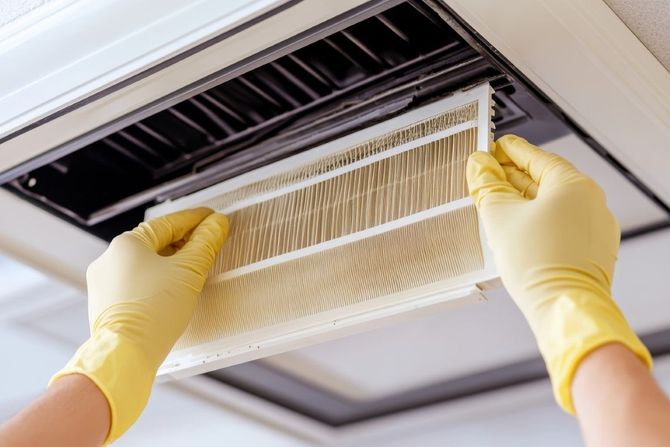 Hands in yellow gloves replacing a dirty HVAC air filter in a ceiling vent