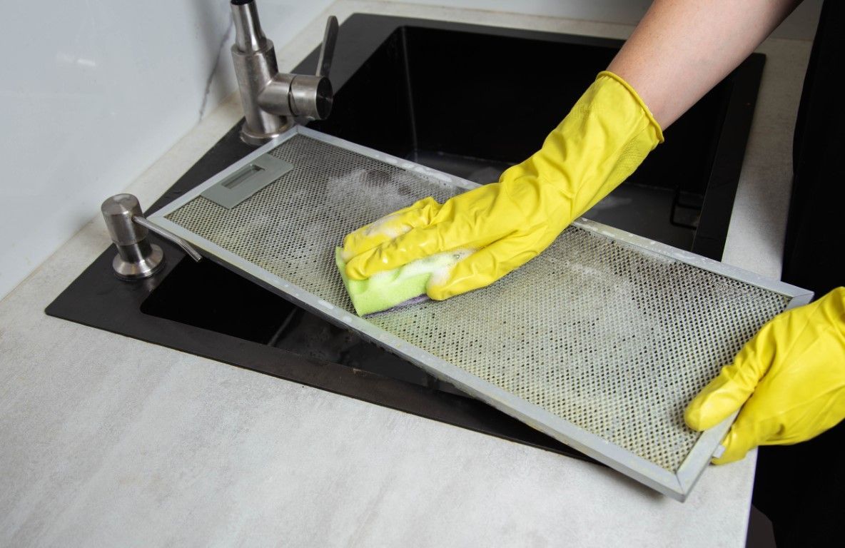 Gloved hands cleaning a metal kitchen filter over a sink with a sponge