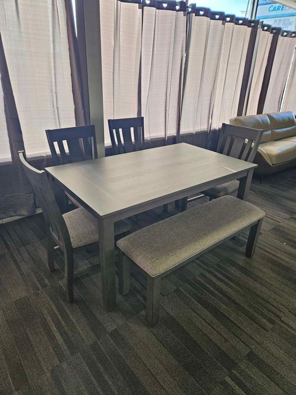 Gray dining table with four chairs and a bench in a room with blinds and wood flooring.