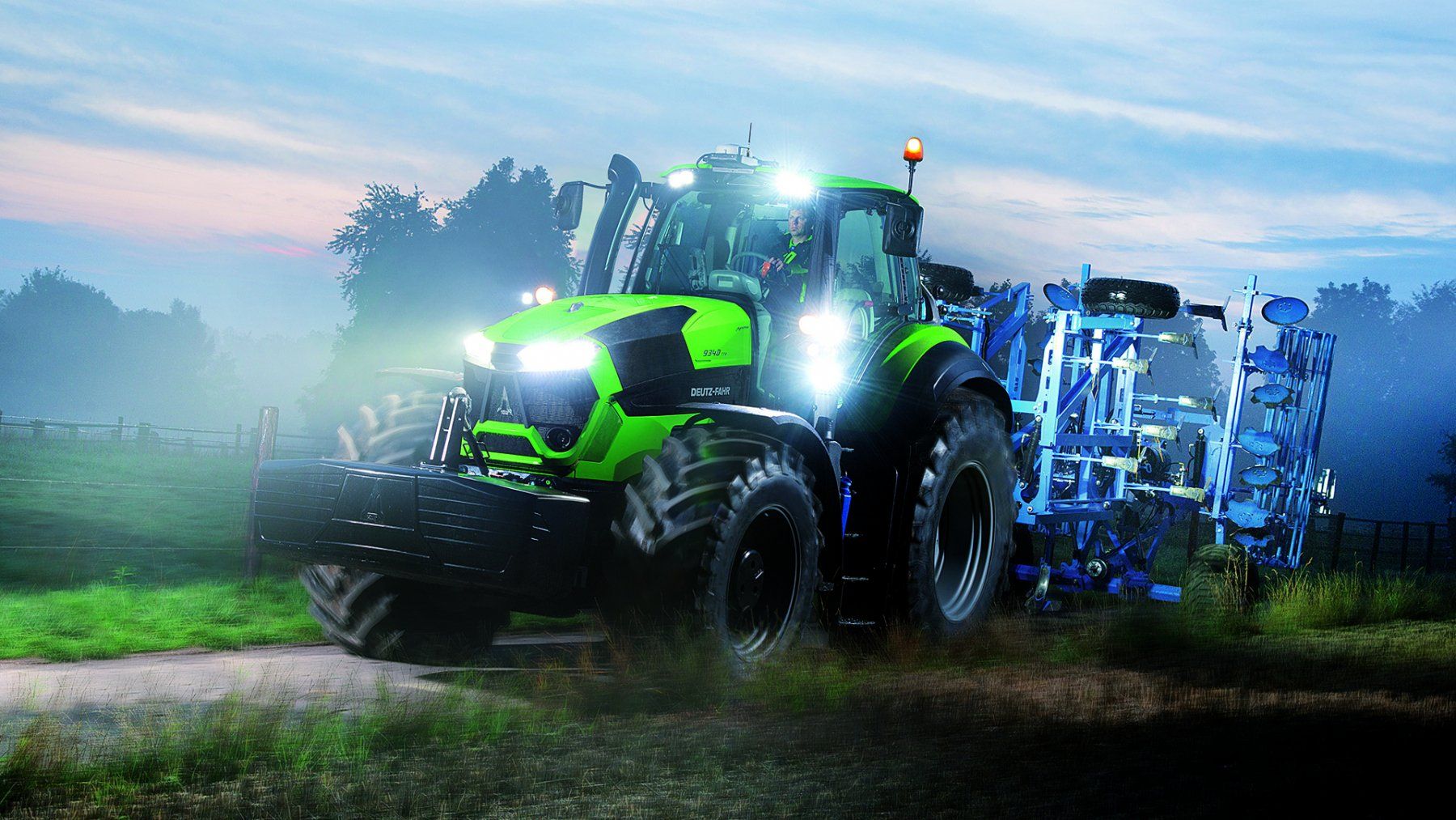 Green tractor pulling blue farm equipment, illuminated at dusk.