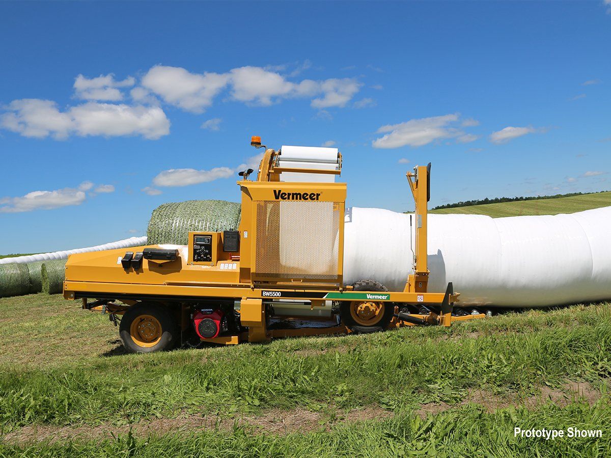 Yellow Vermeer bale wrapper in a field, wrapping a large white plastic tube around hay bales under a blue sky.