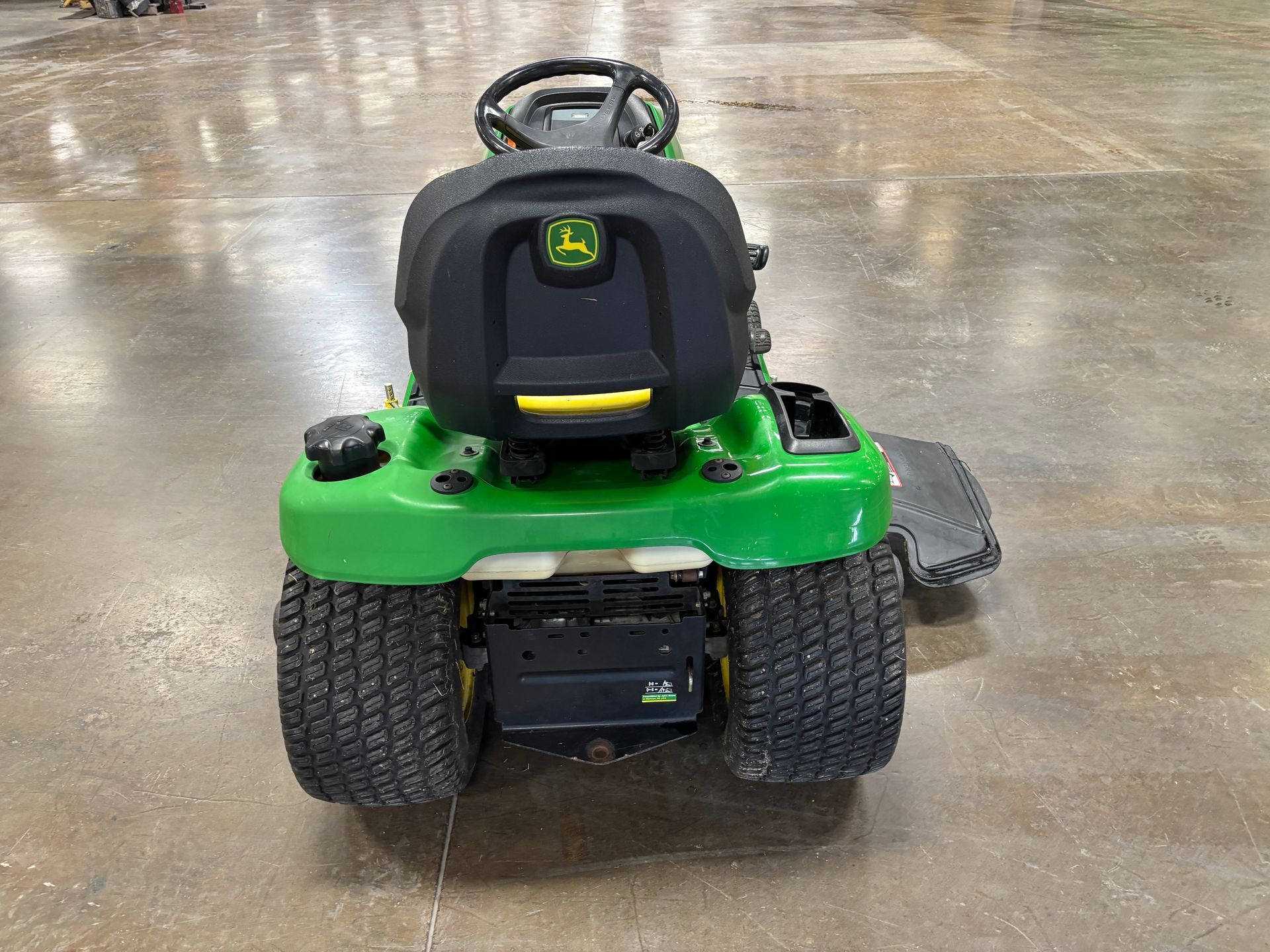 Green and yellow John Deere riding lawn mower, rear view, on a concrete floor.