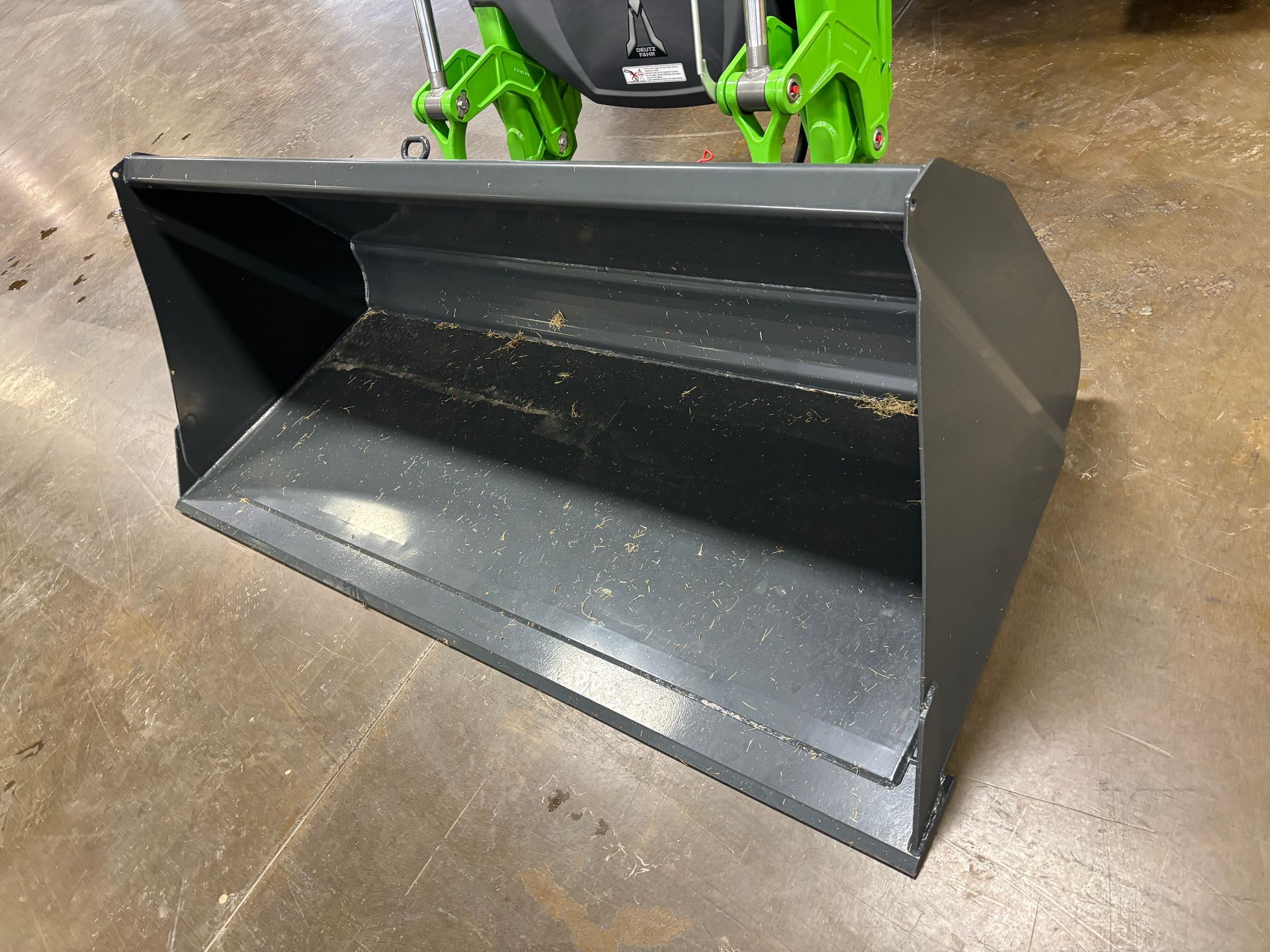 Gray metal bucket attached to a green machine, on a concrete floor.