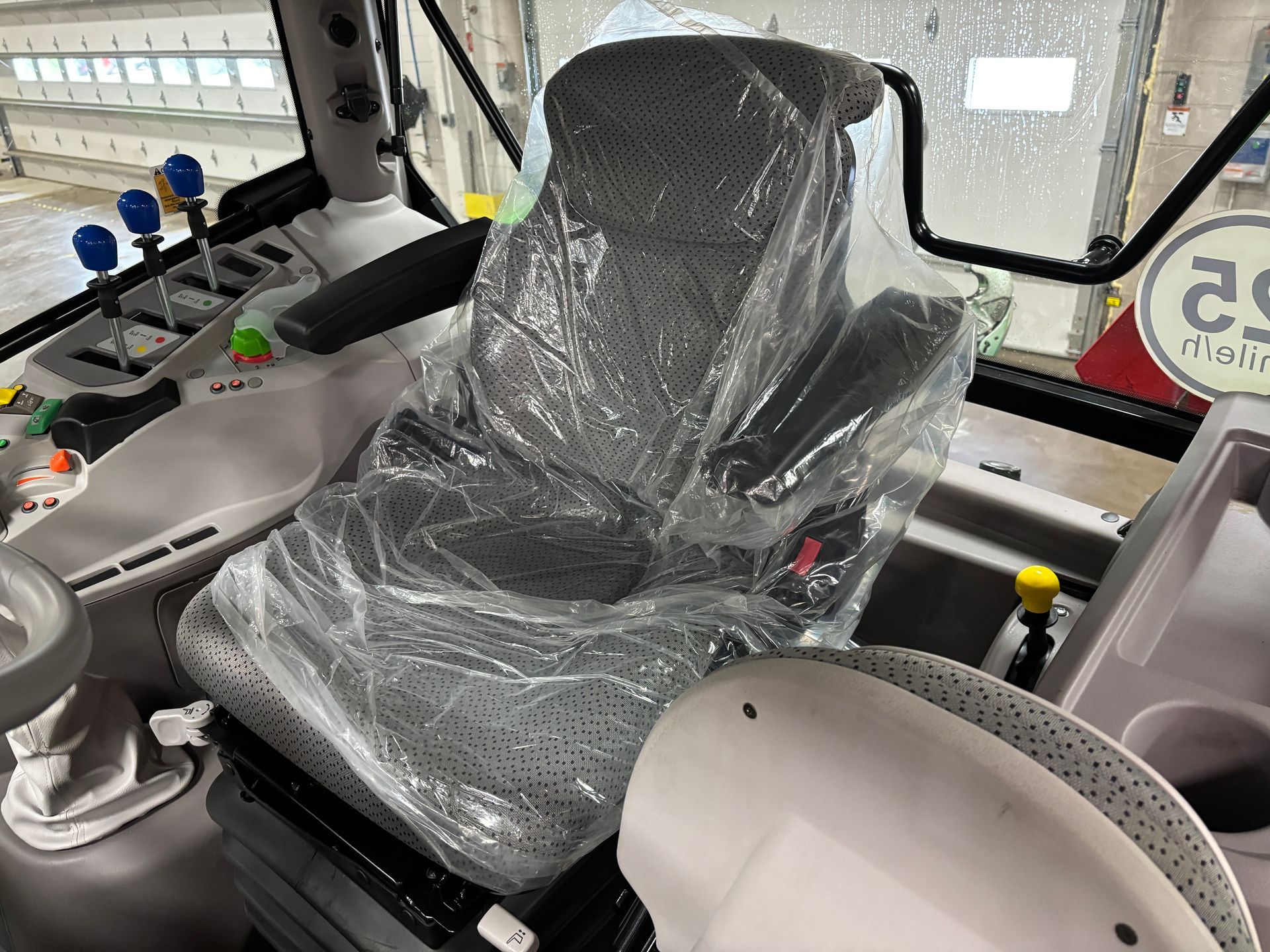 Interior view of tractor cab, focusing on the driver's seat covered in plastic wrap. Gray and white interior with controls visible.