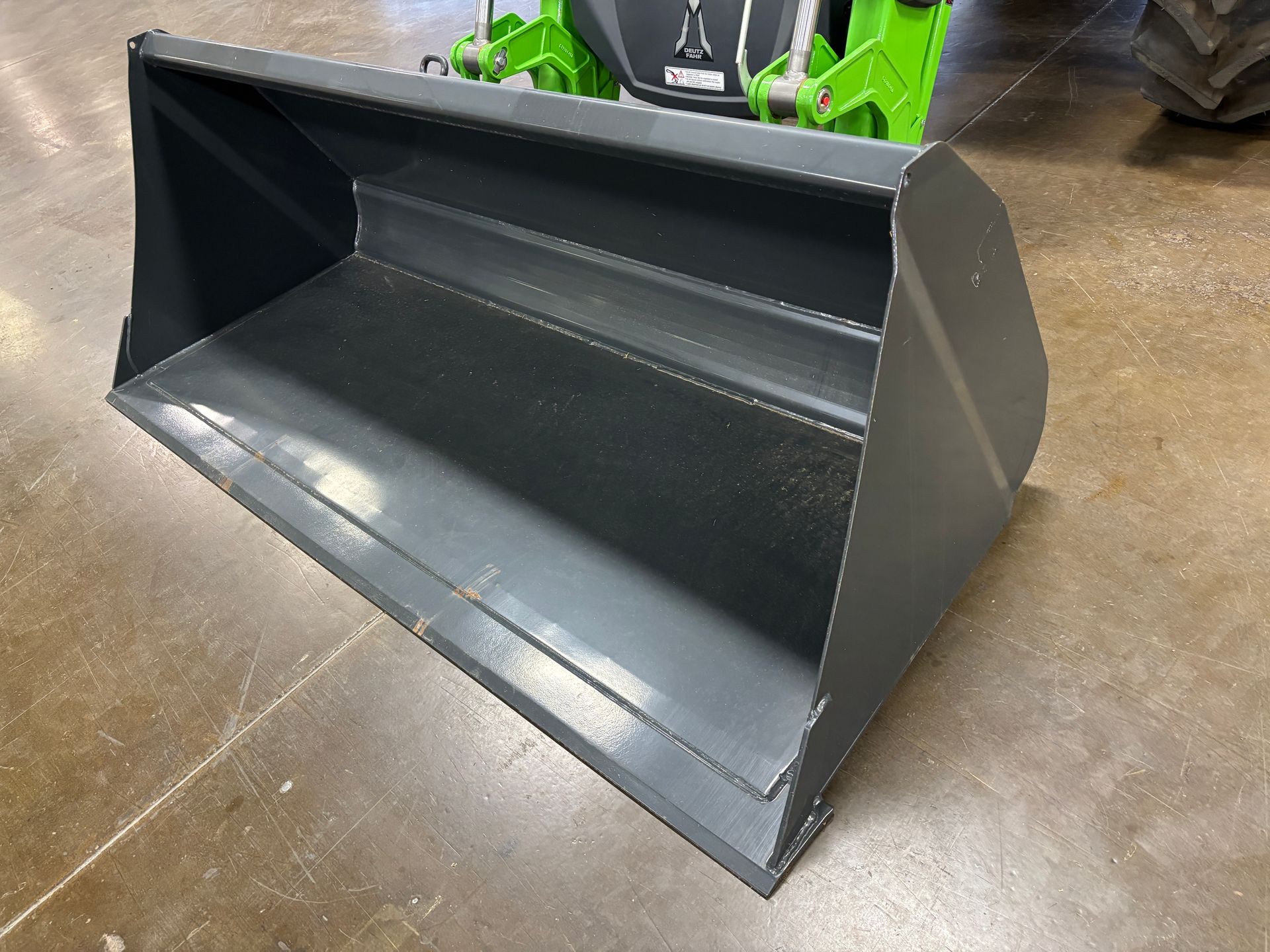 Gray tractor bucket on a concrete floor, with a green tractor in the background.