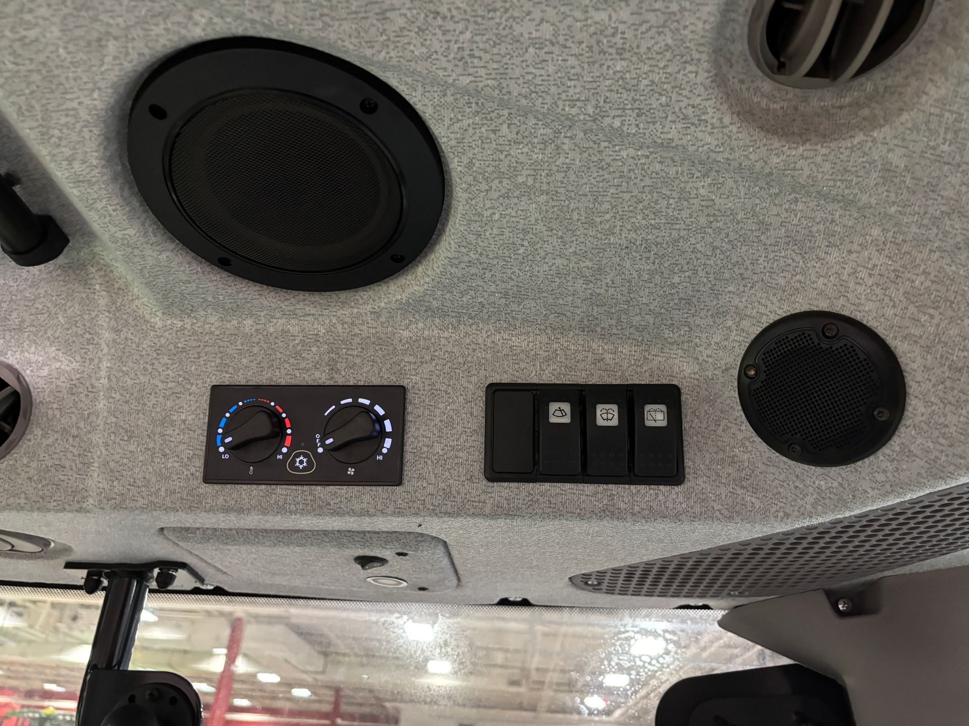 Interior view of vehicle roof. Features speakers, climate controls, and switches, mounted on gray fabric.