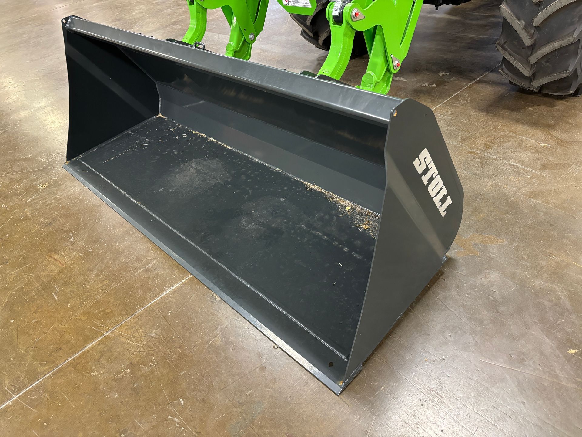Gray Stoll tractor bucket on a tan floor, with green tractor visible in the background.